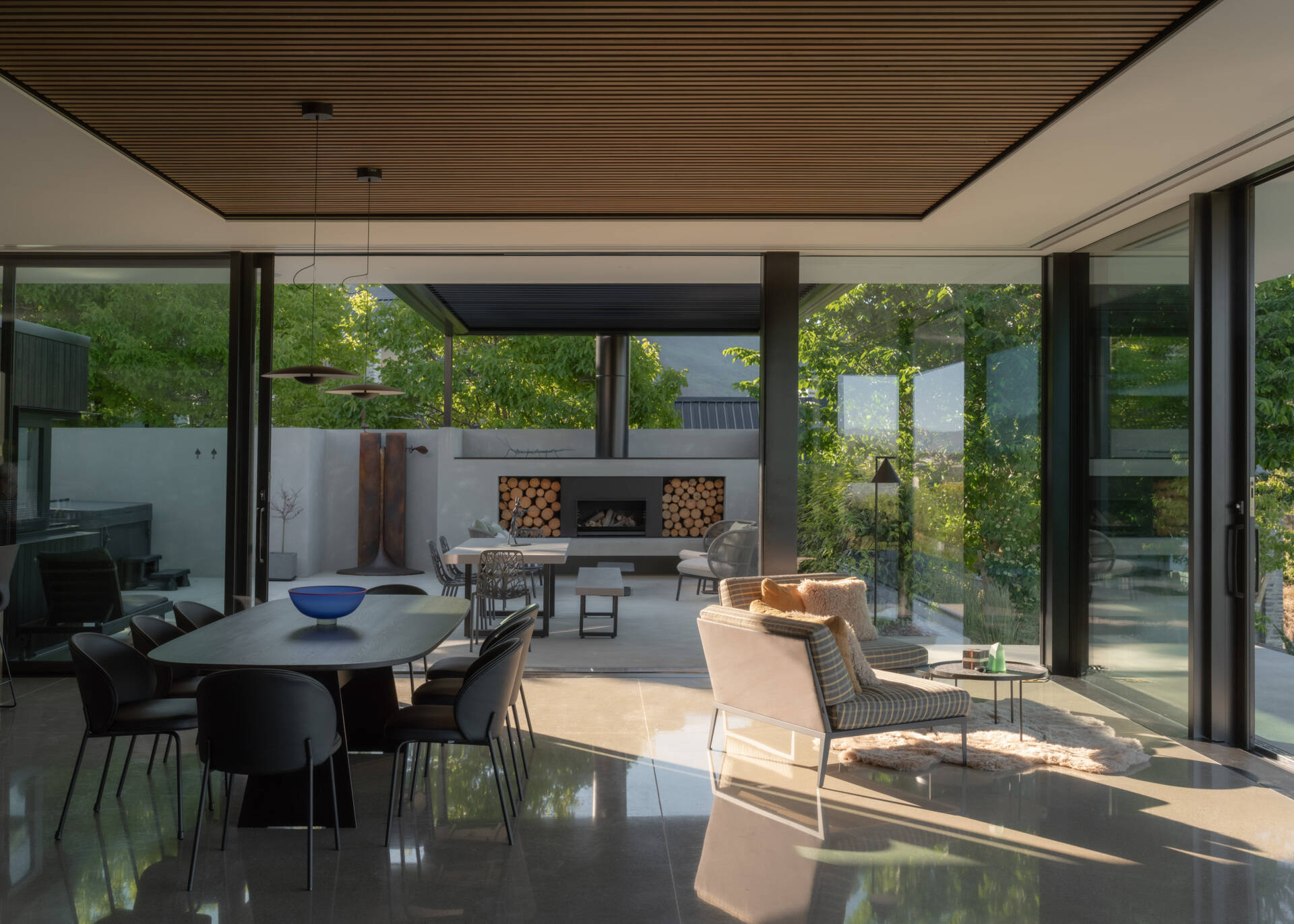 A covered outdoor dining area featuring a built-in fireplace, stacked firewood storage, contemporary outdoor furniture, and large sliding glass doors connecting to the home's interior.