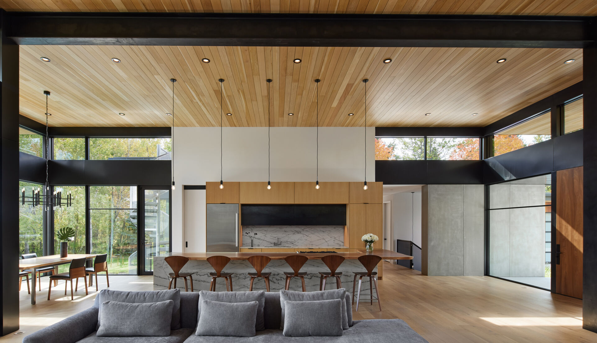 An open-plan modern living room and kitchen with a wood ceiling, exposed black steel beams, pale wood floors, and a large marble-look kitchen island. Tall windows bring natural light into the connected spaces.