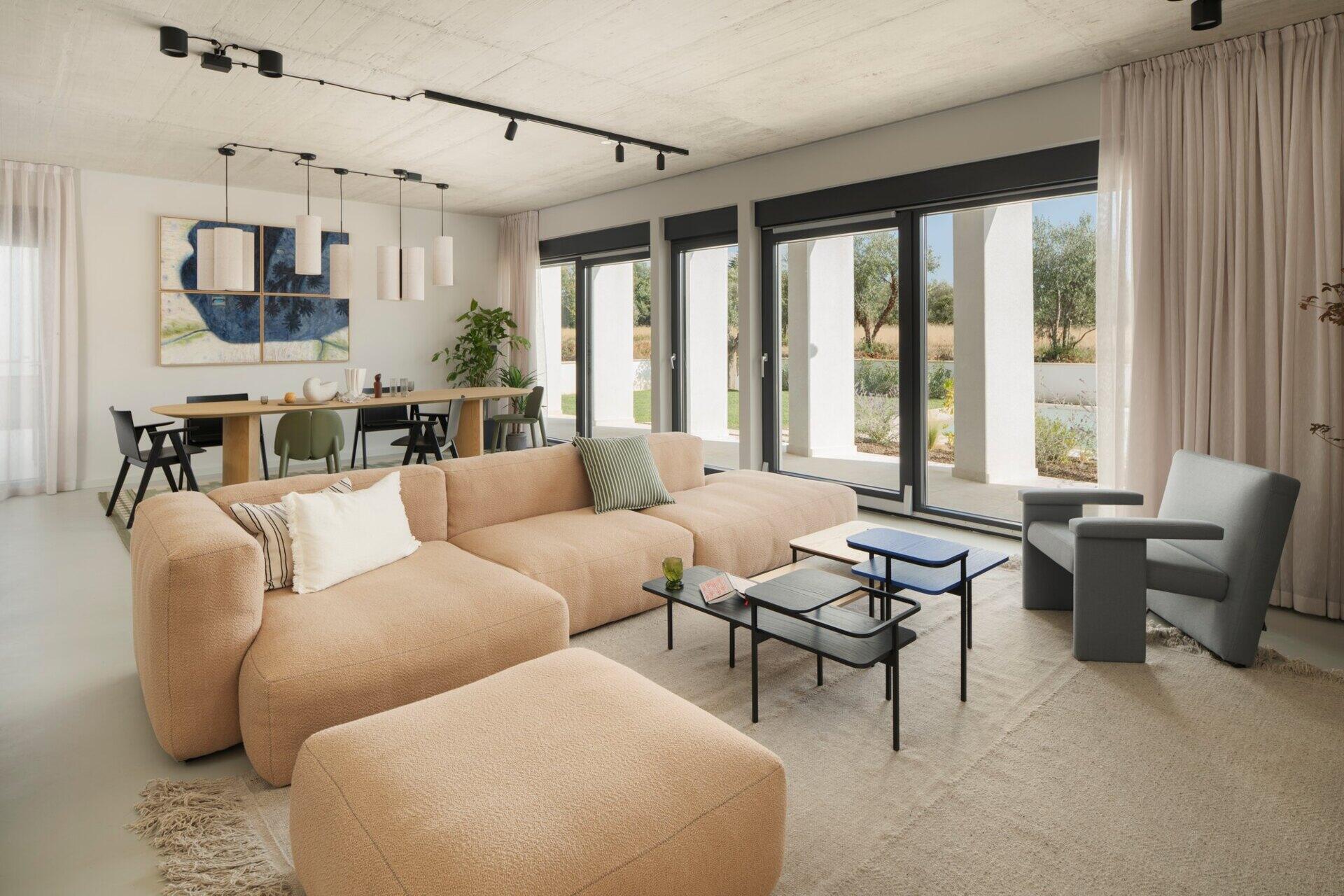 Explore the living room of Arc House in Istria, featuring a peach modular sofa, blue-grey lounge chair, modern coffee tables, exposed concrete ceiling, sheer curtains and warm contemporary interior styling by Jana Piacun.