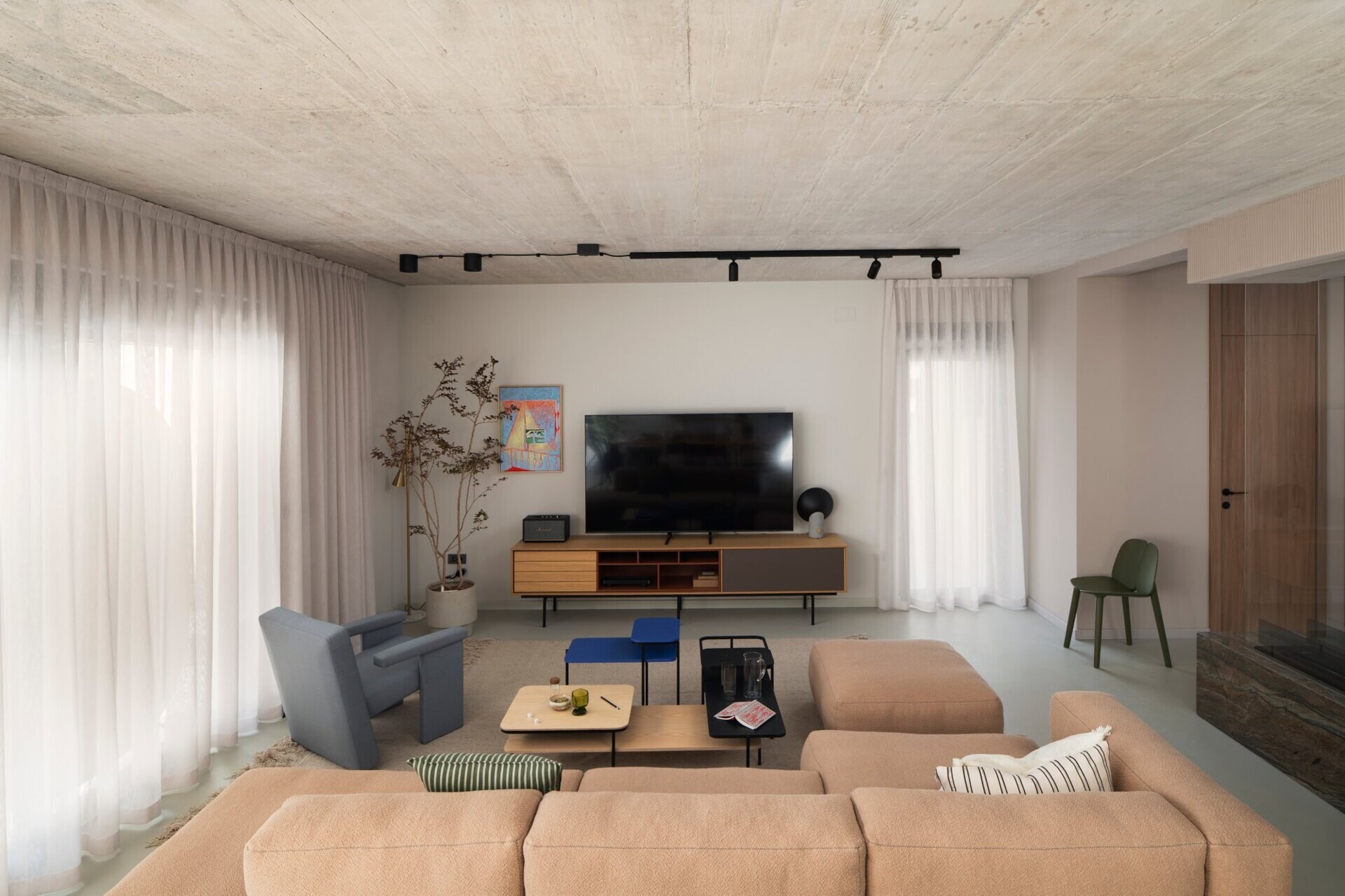 Explore the living room of Arc House in Istria, featuring a peach modular sofa, blue-grey lounge chair, modern coffee tables, exposed concrete ceiling, sheer curtains and warm contemporary interior styling by Jana Piacun.