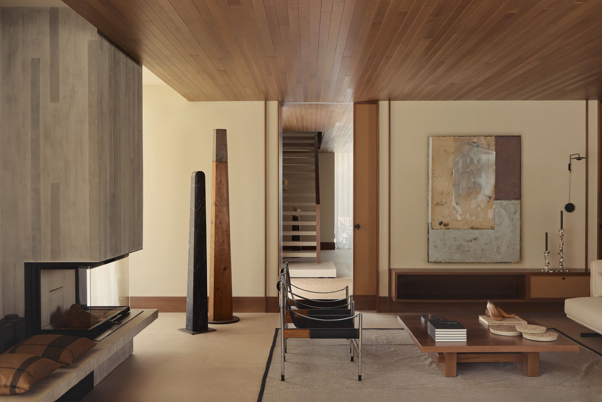 Explore a modern living room in Spain with a sculptural concrete fireplace, walnut coffee table, vintage armchairs, contemporary artwork, and warm wood ceiling.