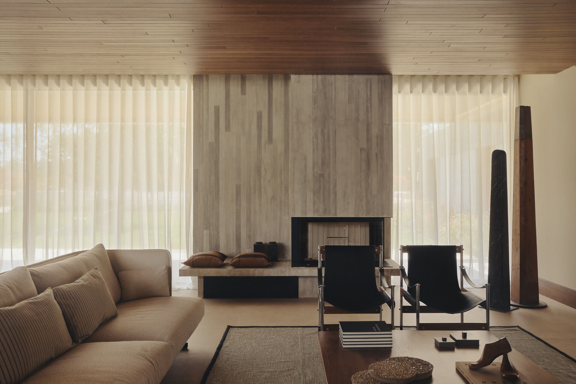 Explore a modern living room in Spain with a sculptural concrete fireplace, walnut coffee table, vintage armchairs, contemporary artwork, and warm wood ceiling.