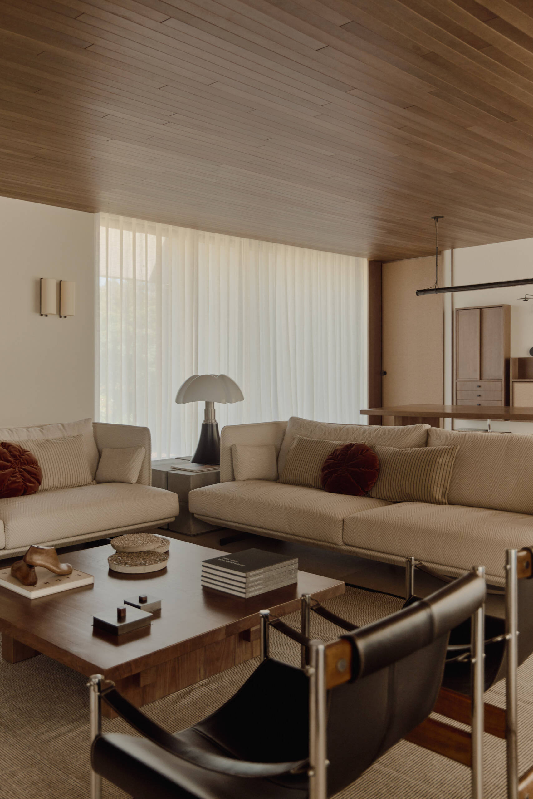 Explore a modern living room in Spain with a sculptural concrete fireplace, walnut coffee table, vintage armchairs, contemporary artwork, and warm wood ceiling.