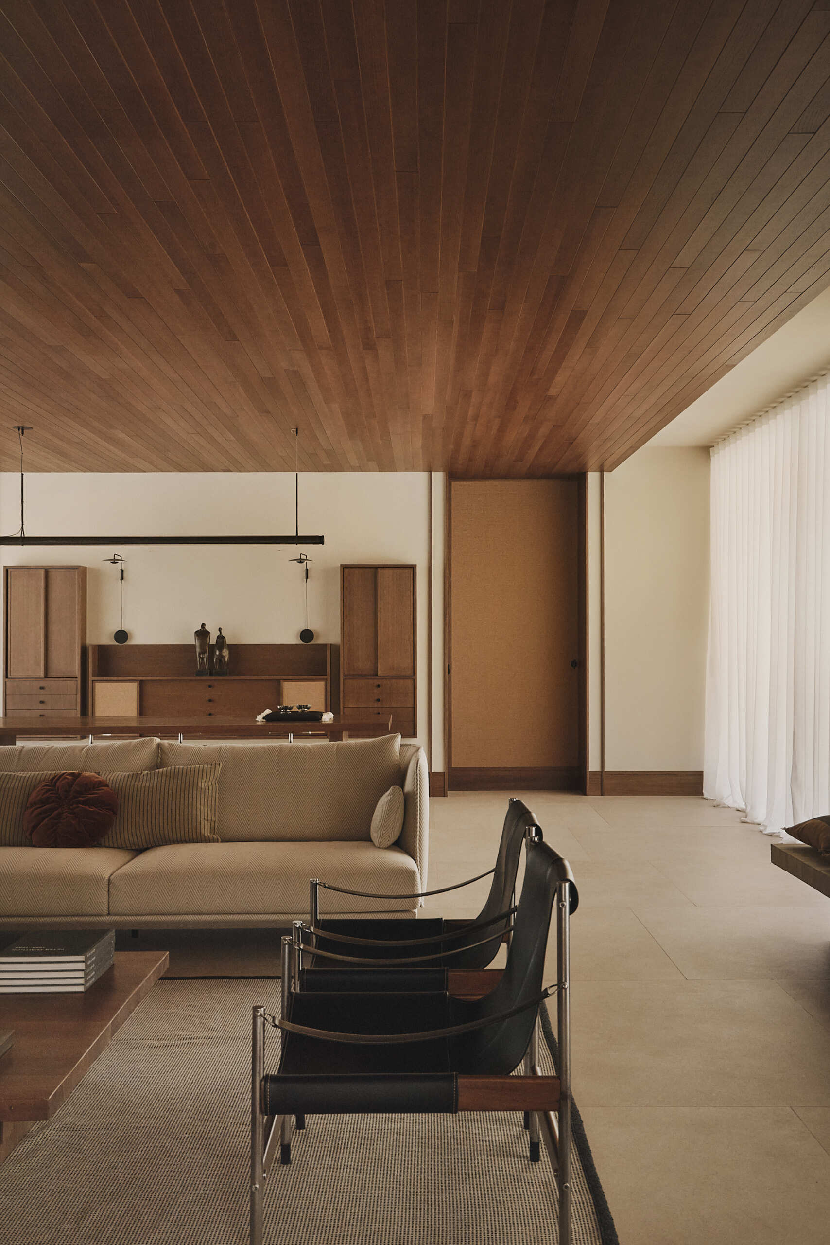 Explore a modern living room in Spain with a sculptural concrete fireplace, walnut coffee table, vintage armchairs, contemporary artwork, and warm wood ceiling.