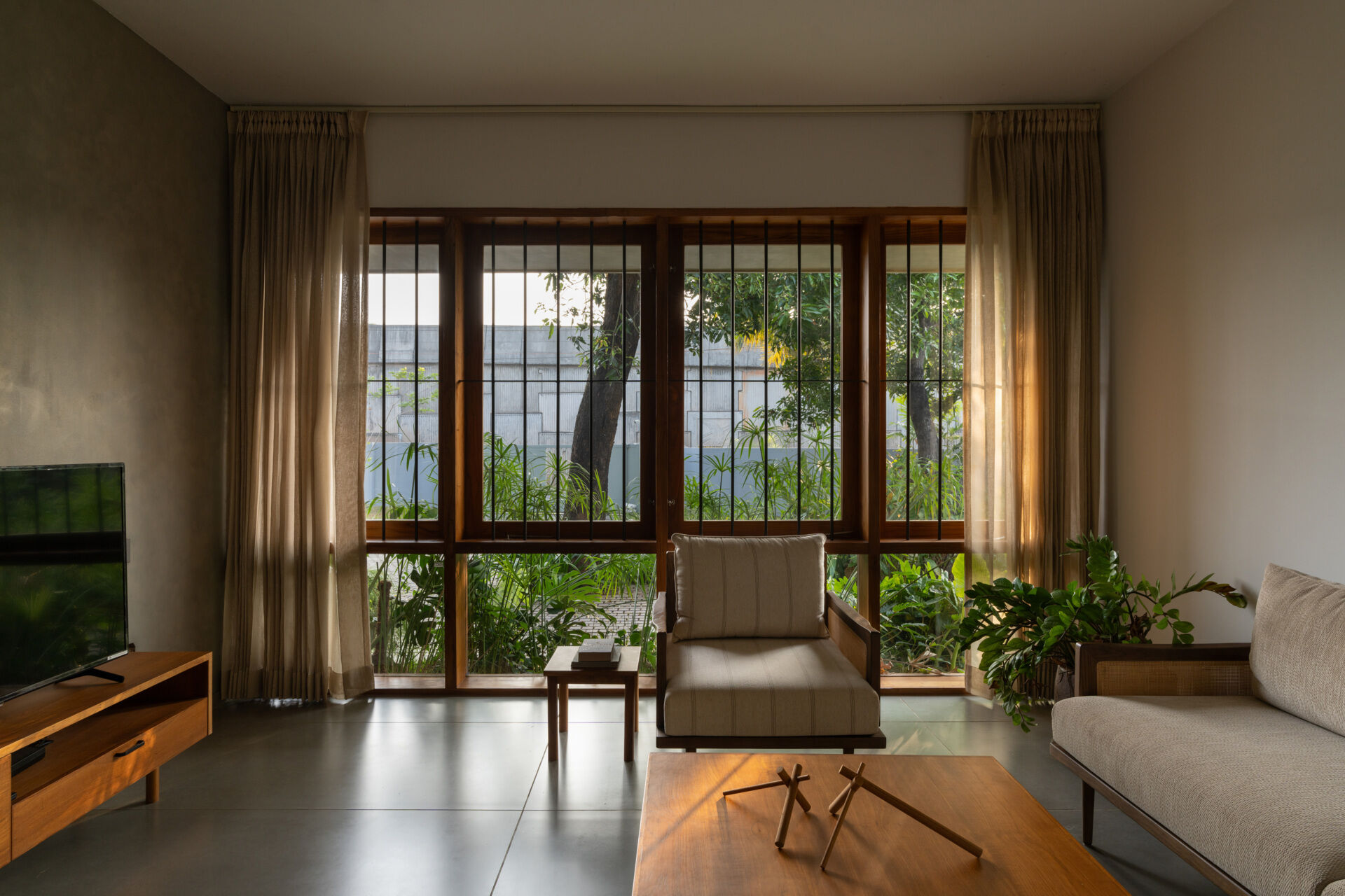 Discover a contemporary living room with large teak-framed windows, neutral furniture, wooden accents, and views of a lush tropical garden.