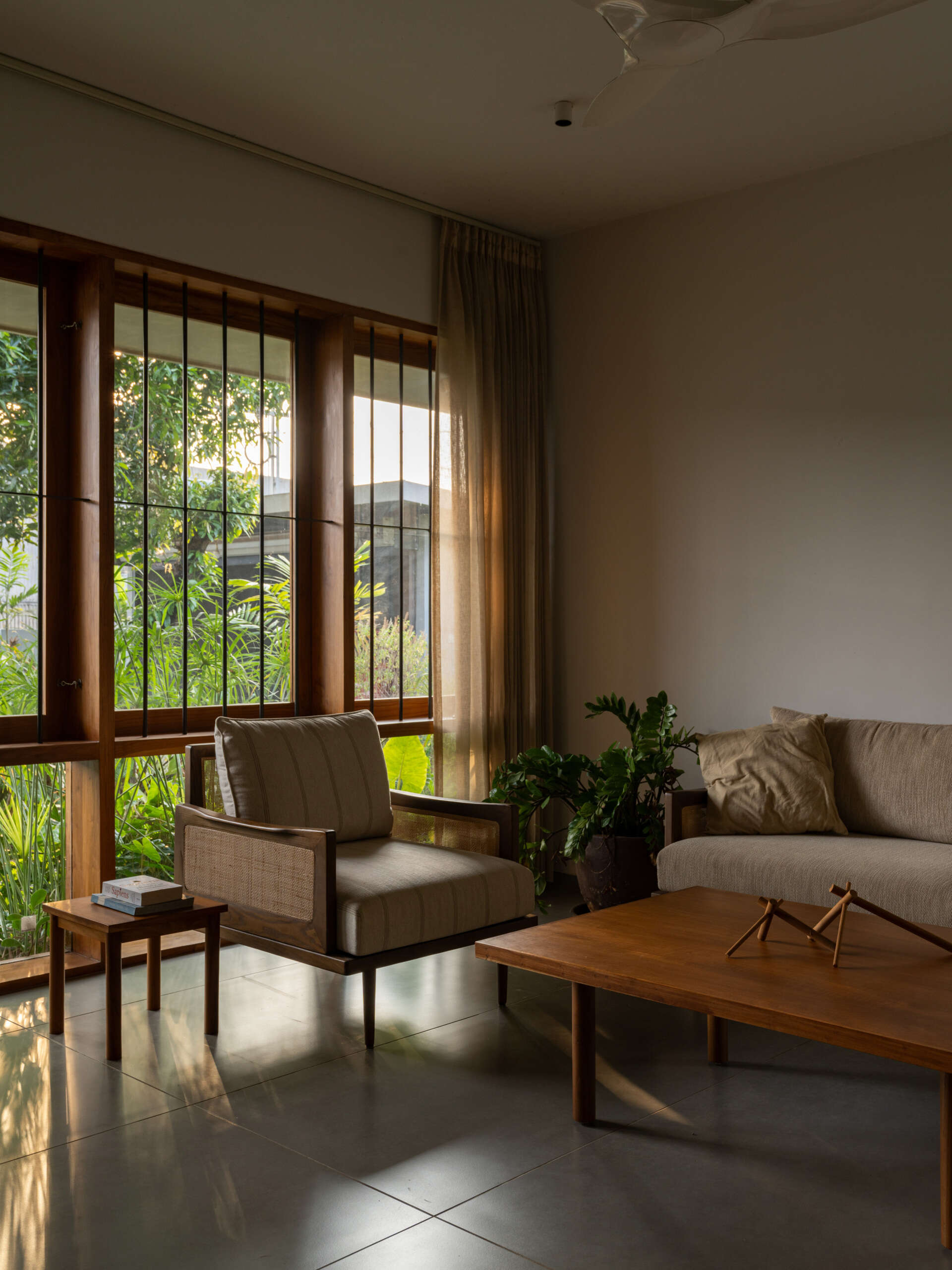 Discover a contemporary living room with large teak-framed windows, neutral furniture, wooden accents, and views of a lush tropical garden.