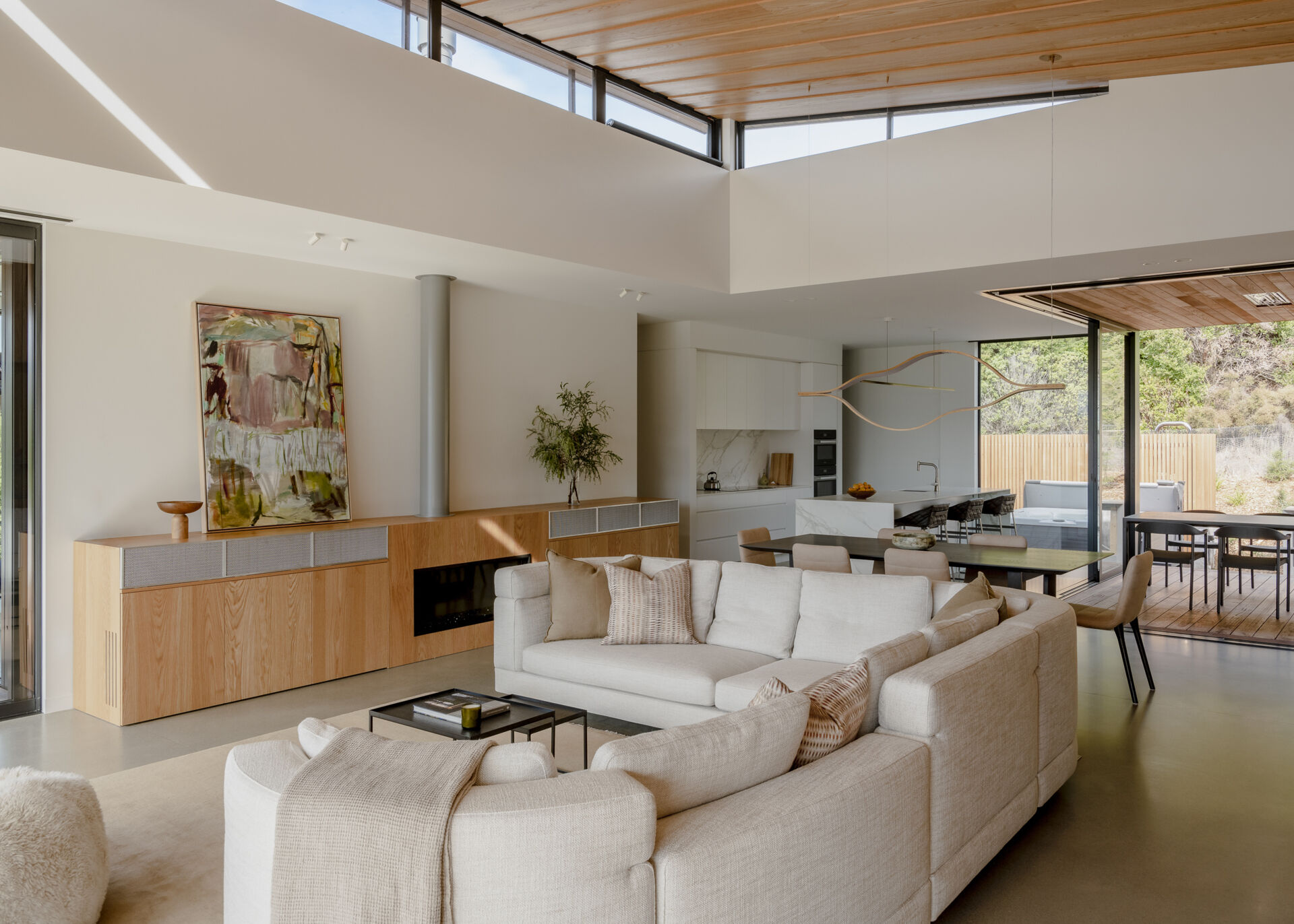 Open-plan living room featuring a cream sectional sofa, built-in wood cabinetry, modern fireplace, and white kitchen with a marble island. High windows and a wood-lined ceiling bring natural light and warmth to the interior.