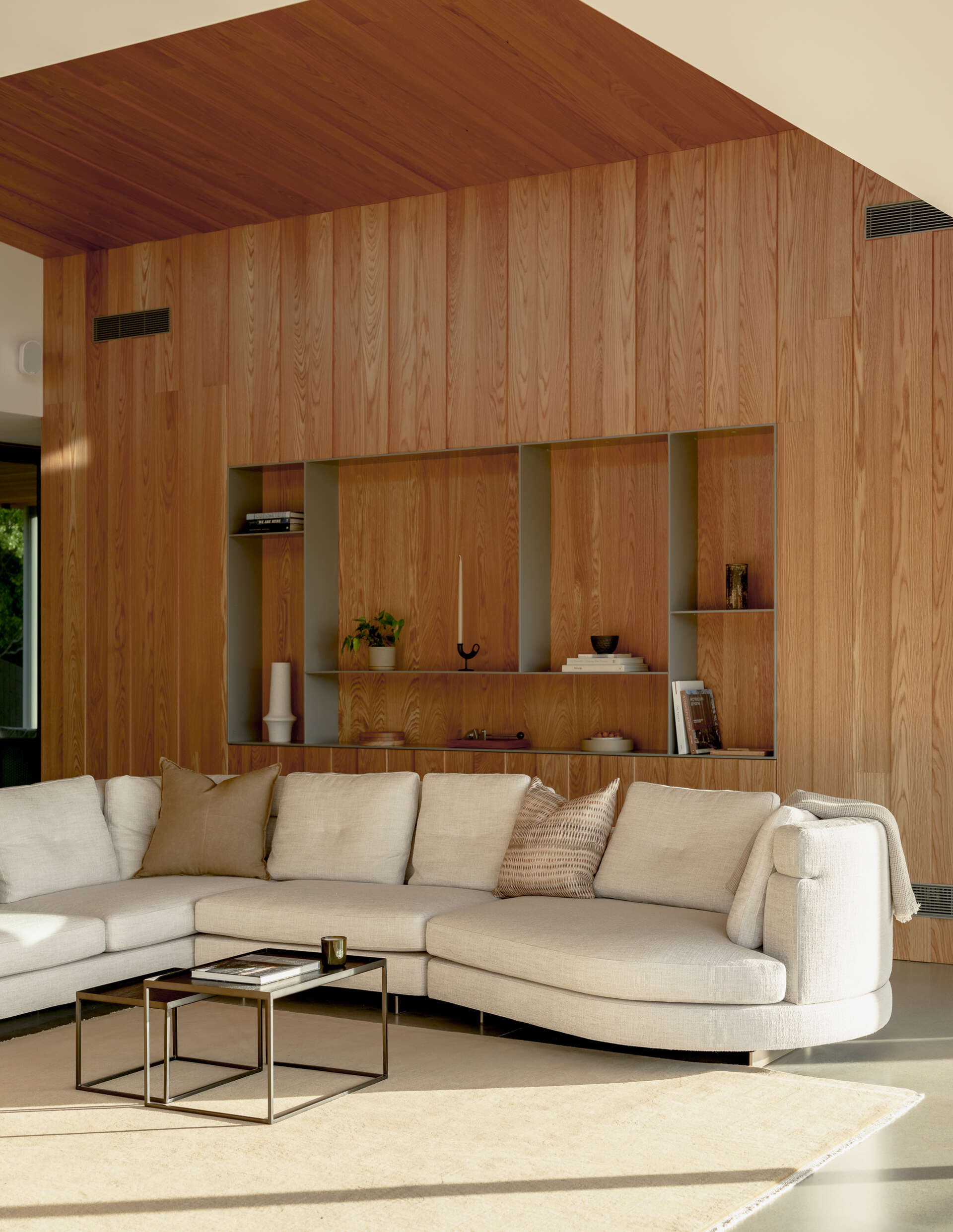 Warm minimalist living room featuring a floor-to-ceiling wood feature wall with recessed display shelves, a curved cream sectional sofa, and black nesting coffee tables. Natural wood grain adds texture to the contemporary interior.
