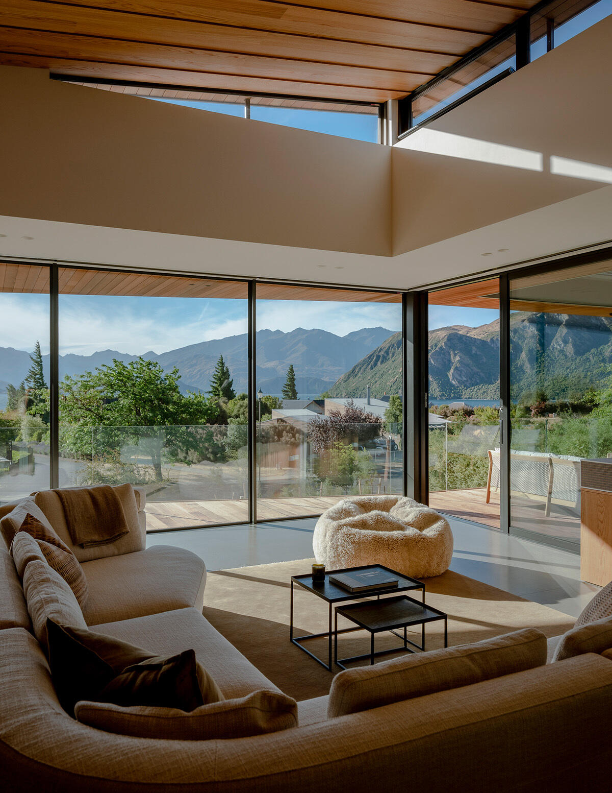 Modern living room with a large cream sectional sofa, floor-to-ceiling corner windows, warm wood ceiling, and panoramic mountain views. High windows beneath the angled roof bring additional sunlight into the space.