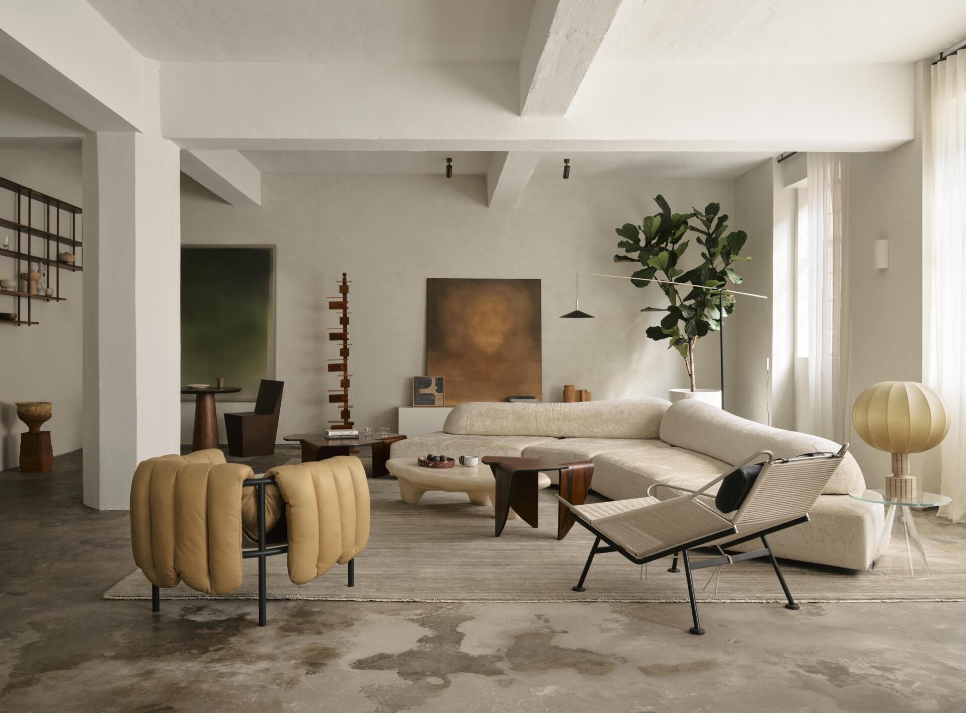 A spacious contemporary living room with a curved cream sectional sofa, sculptural accent chairs, low coffee tables, and a neutral area rug. White-painted concrete ceiling beams, large windows, and a potted tree complete the industrial apartment interior.