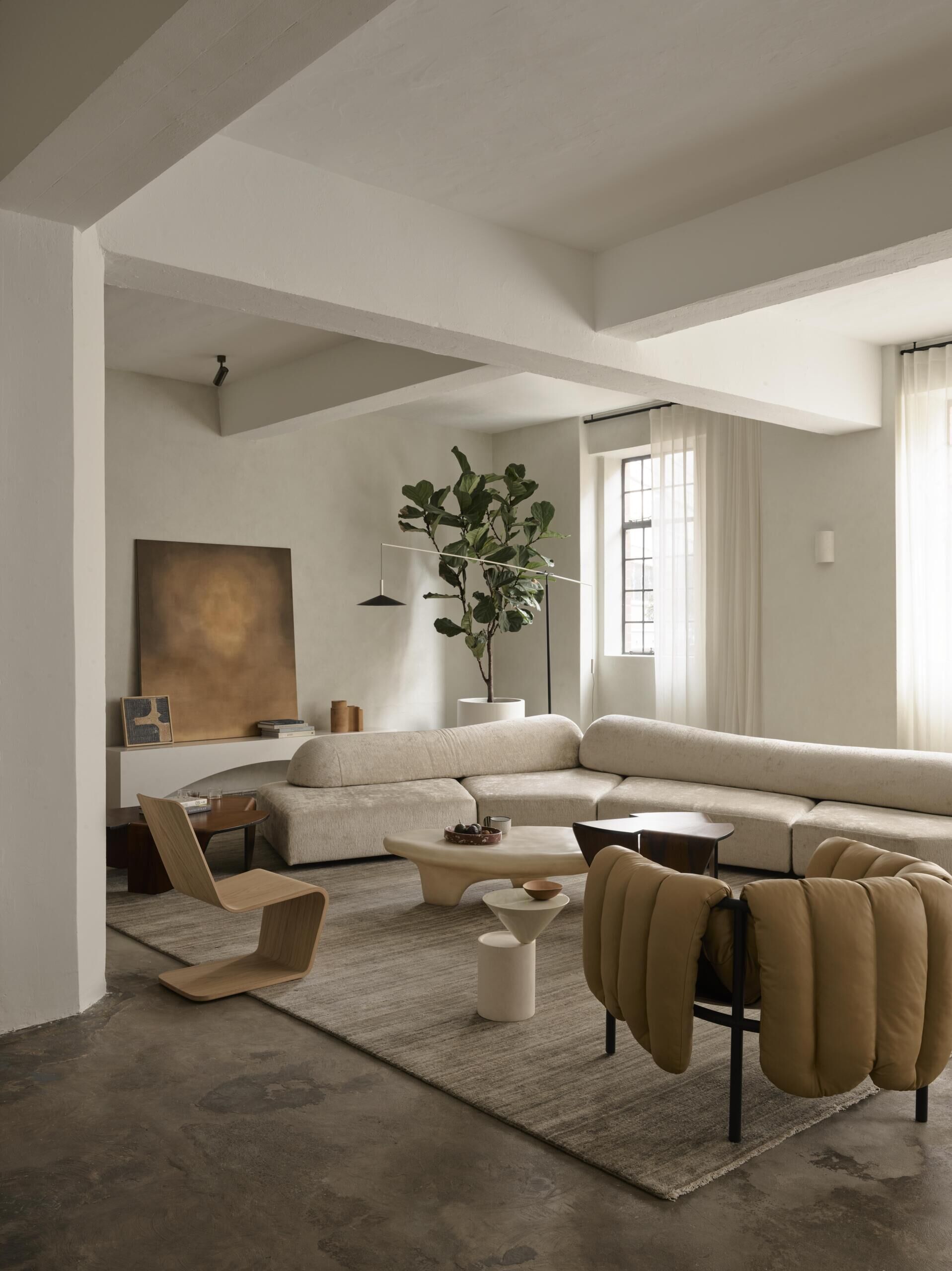A minimalist living room with an oversized curved sofa, sculptural coffee tables, upholstered accent chair, and floor-to-ceiling sheer curtains. Exposed concrete beams and warm neutral furnishings bring together industrial architecture and contemporary interior design.