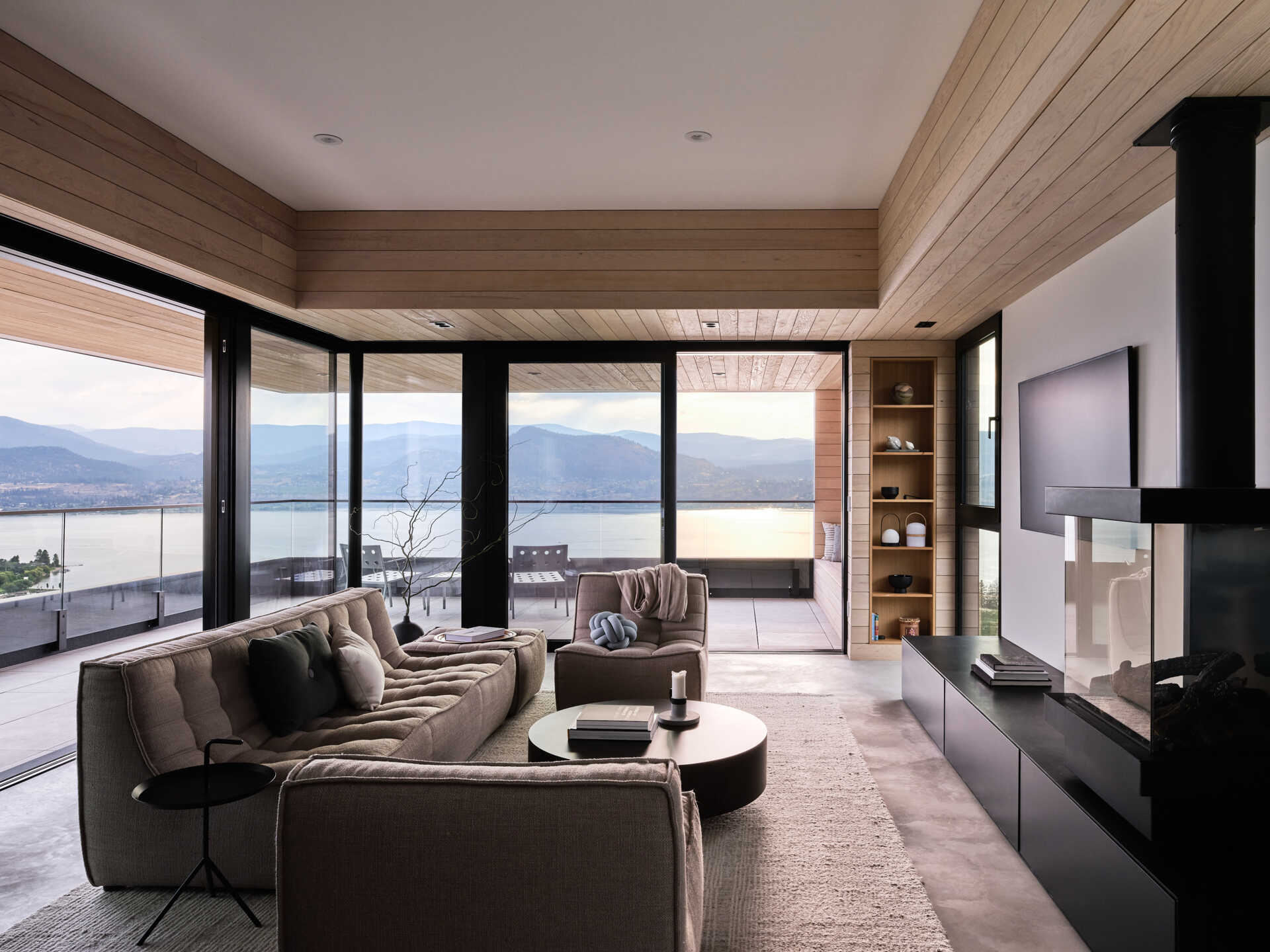 A modern living room with a neutral sectional sofa, black suspended fireplace, and floor-to-ceiling glass walls overlooking Okanagan Lake. Sliding doors open to a covered terrace with glass railings and mountain views.