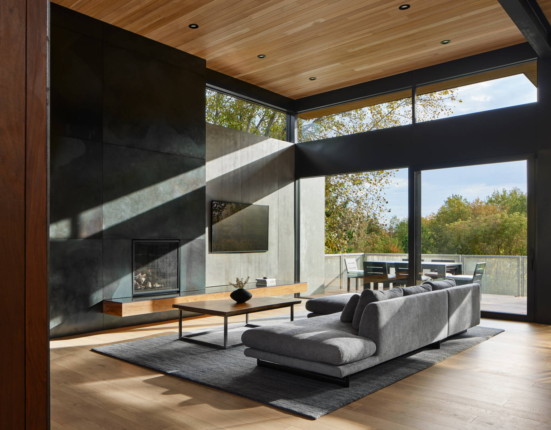 A contemporary living room with a gray sofa, dark fireplace wall, wood ceiling, and expansive corner windows. Sunlight enters through the glass, with an outdoor dining deck beyond.