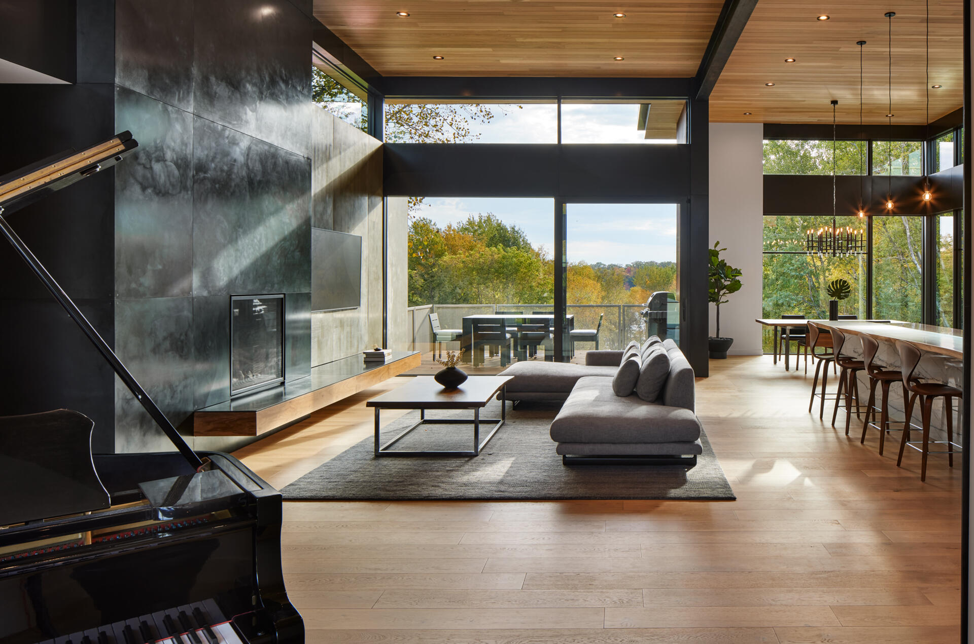 A modern living room with a tall dark fireplace wall, gray sectional sofa, pale wood flooring, and a wood ceiling. Large glass doors open onto an outdoor deck overlooking the trees.