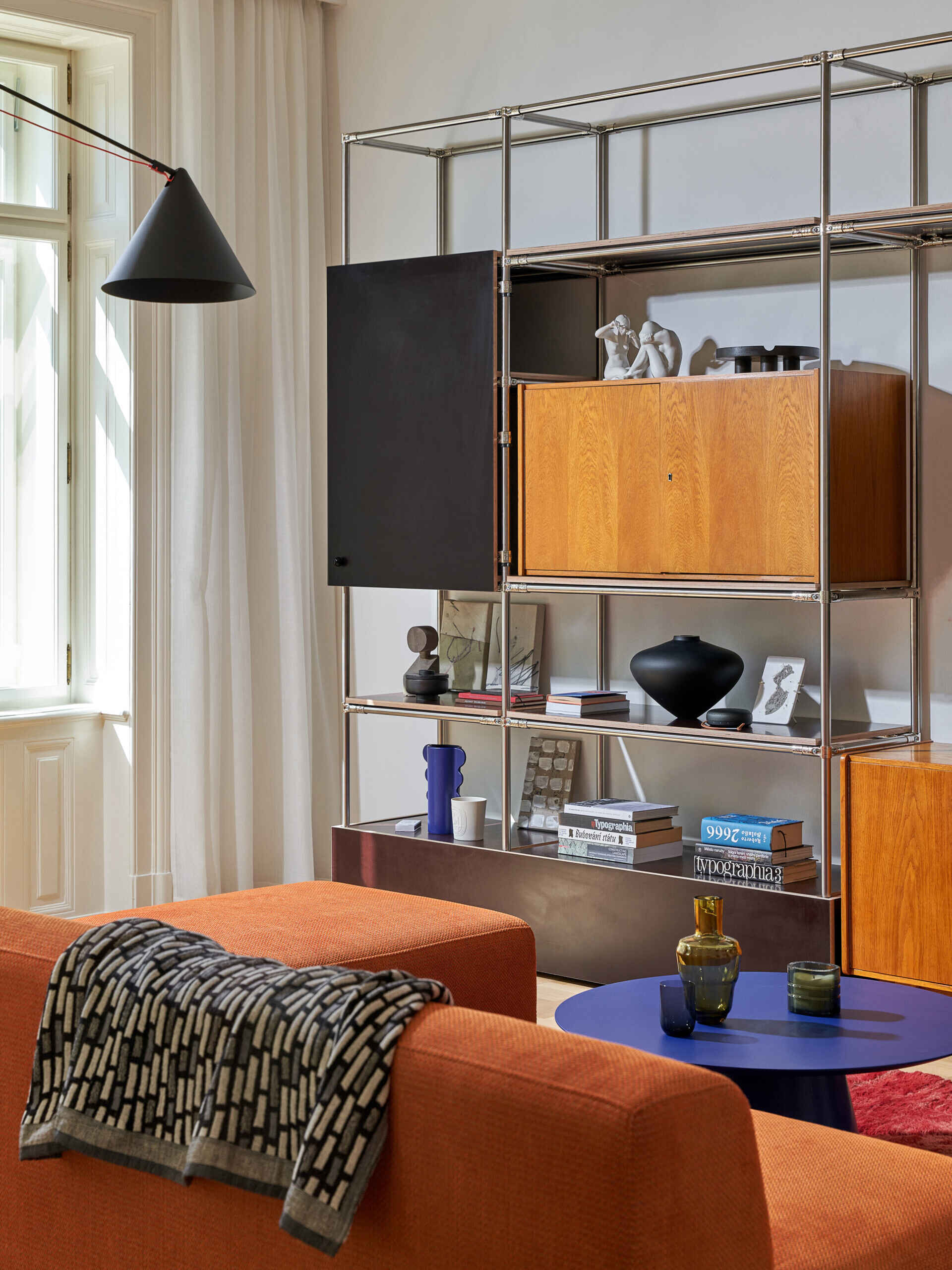 An orange sectional sofa anchors this contemporary living room, accompanied by a blue circular coffee table and open metal shelving incorporating warm-toned cabinets and decorative objects.
