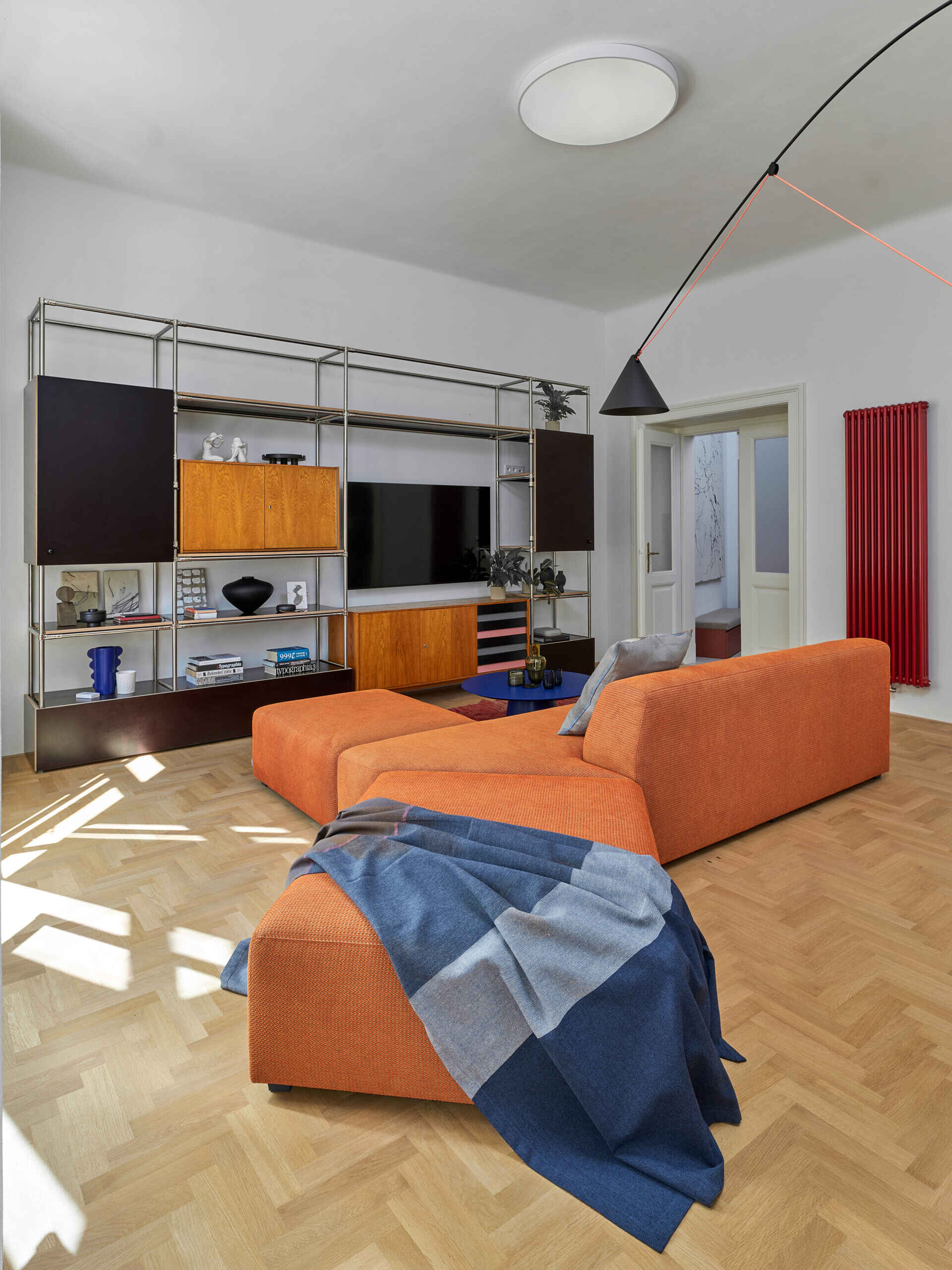 A colorful modern living room with a large orange sectional sofa, blue coffee table, and floor-to-ceiling metal shelving. Original panelled doors and herringbone flooring add historic character.