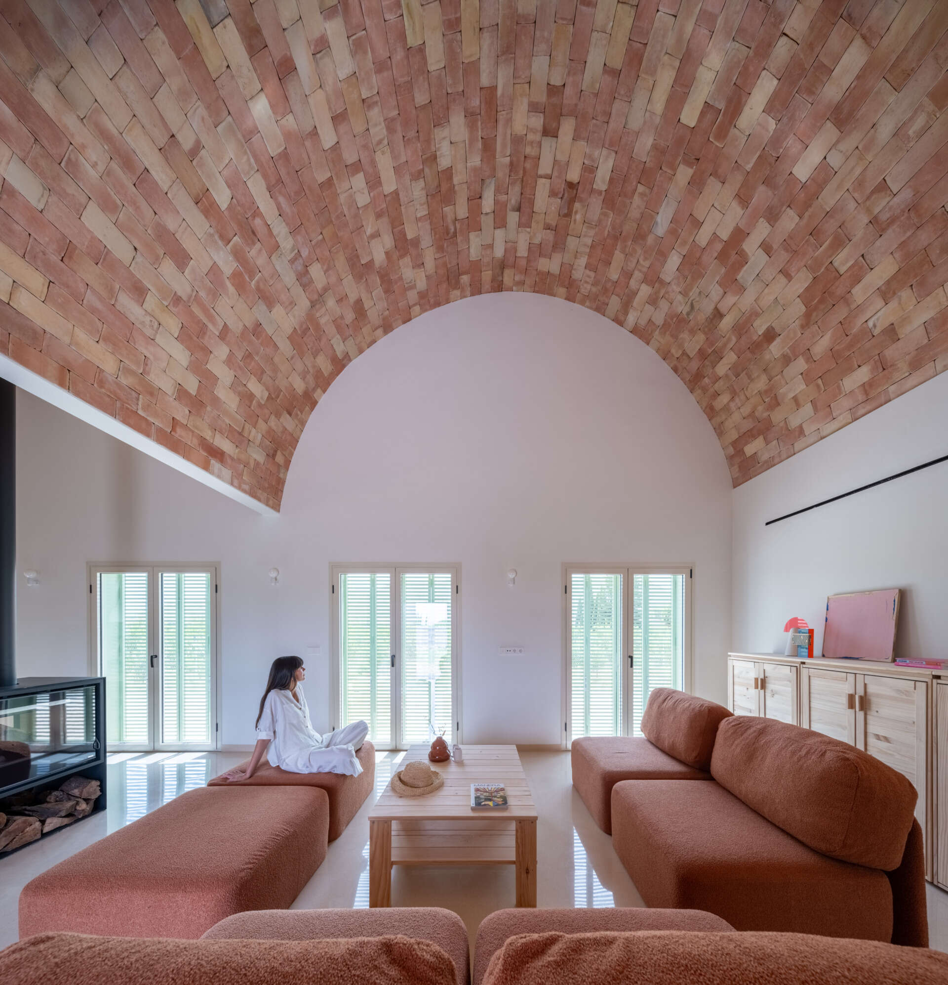 Step inside the living room of Casa Vereda in Spain, where a dramatic curved brick ceiling rises above rust-coloured modular sofas, terrazzo flooring, and contemporary timber furniture. A beautiful combination of traditional Catalan architecture and modern interior design.
