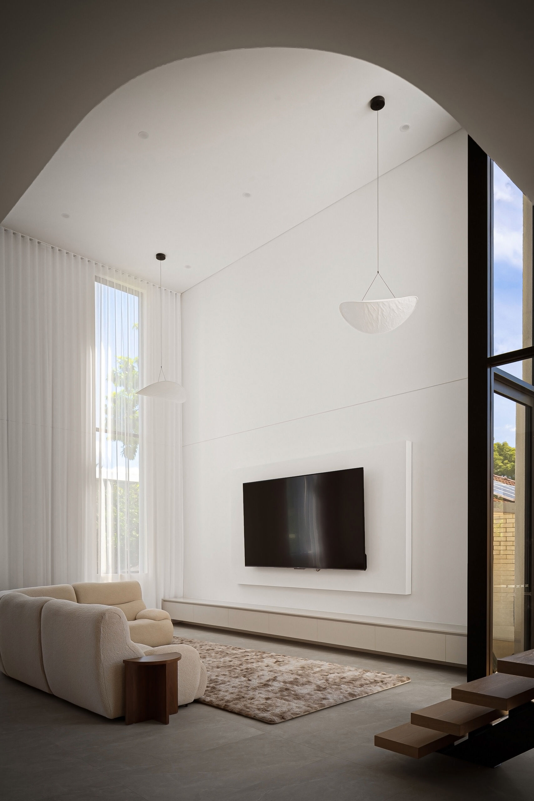A modern double-height living room with tall windows, floor-to-ceiling curtains, and white sculptural pendant lights. Curved ceiling details, pale upholstered seating, a wall-mounted television, and floating wood stair treads complete the interior.