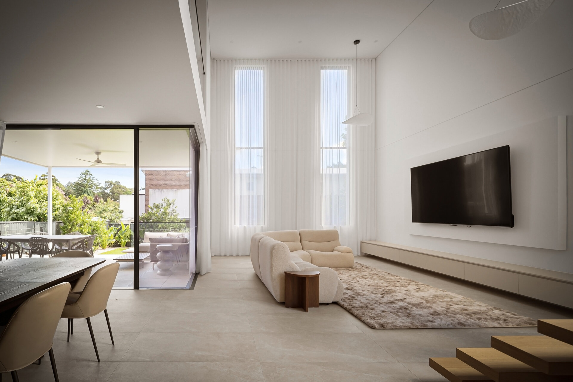 A bright contemporary living room featuring a double-height ceiling, tall narrow windows, and large sliding glass doors opening to a covered outdoor dining area. Pale lounge chairs, a textured rug, and a long floating media cabinet furnish the space.