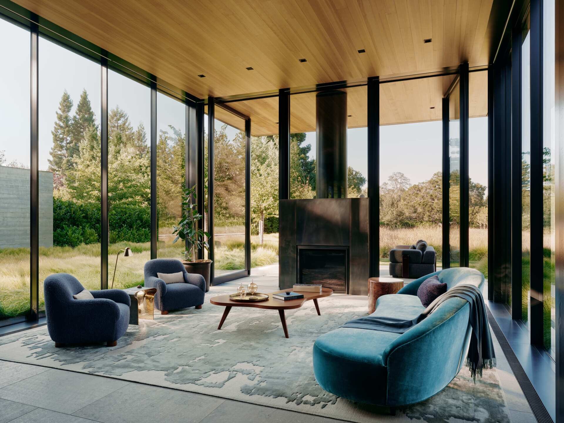  A curved blue-green sofa, rounded lounge chairs, sculptural furniture, and a contemporary black fireplace create a colorful interior surrounded by garden views at this California home.