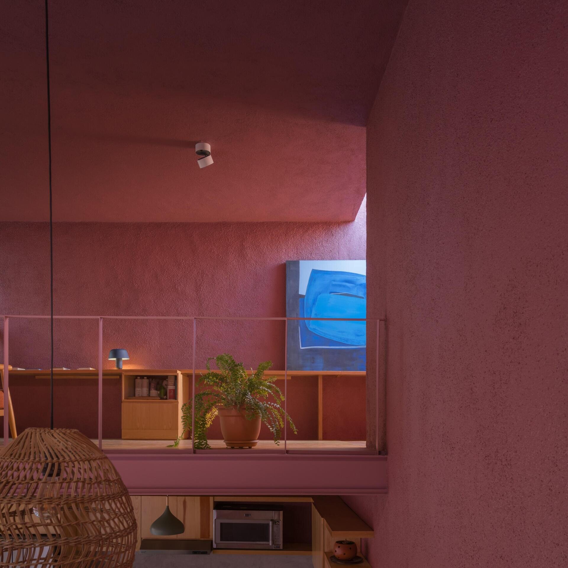 A mezzanine home office in a modern Mexican house features a long built-in wood desk, overhead skylight, pink walls, blue artwork, and a small sitting area overlooking the living room.