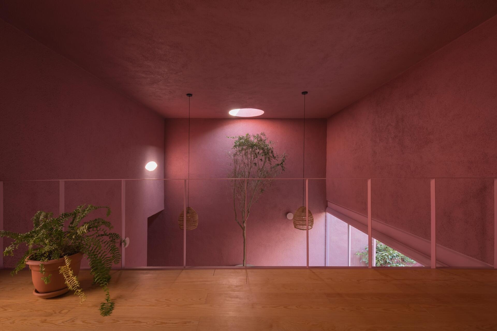A mezzanine home office in a modern Mexican house features a long built-in wood desk, overhead skylight, pink walls, blue artwork, and a small sitting area overlooking the living room.