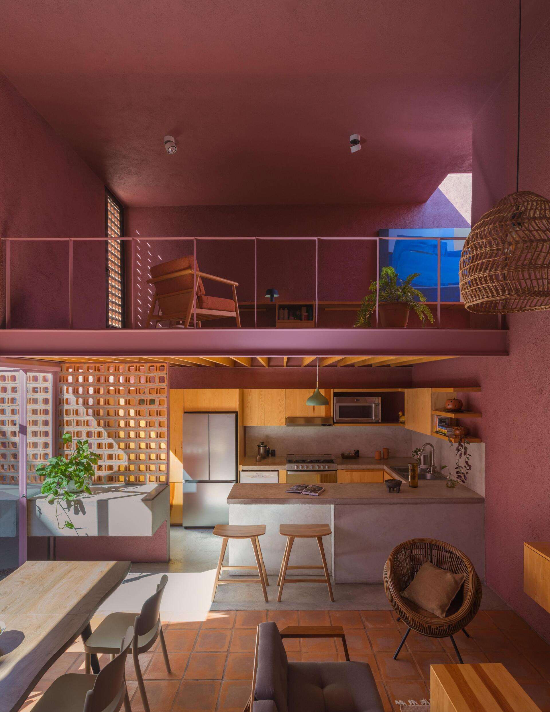 A mezzanine home office in a modern Mexican house features a long built-in wood desk, overhead skylight, pink walls, blue artwork, and a small sitting area overlooking the living room.