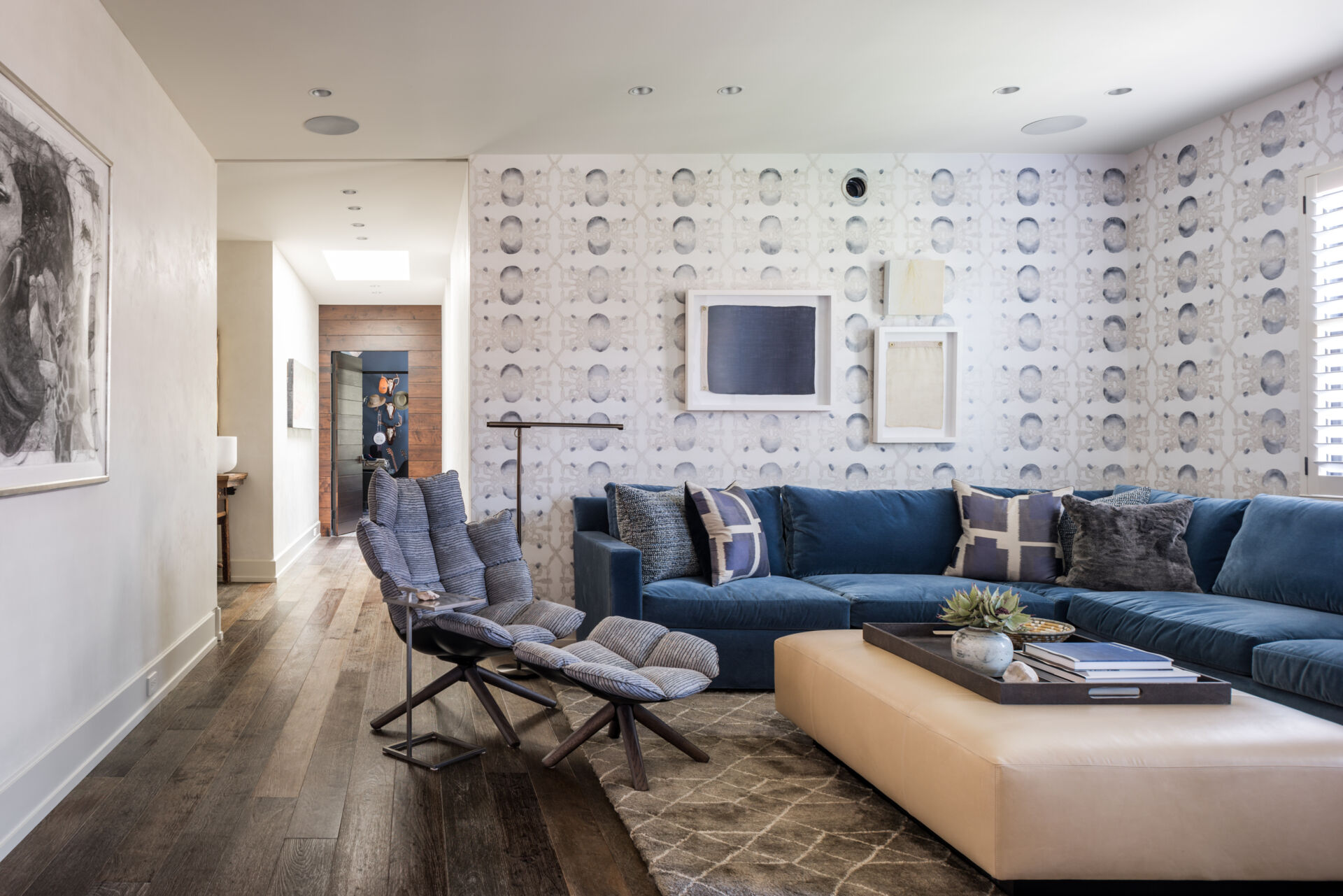 A cozy contemporary media room with a navy-blue sectional sofa, oversized cream ottoman, sculptural lounge chair, blue-gray patterned wallpaper, and textured furnishings.
