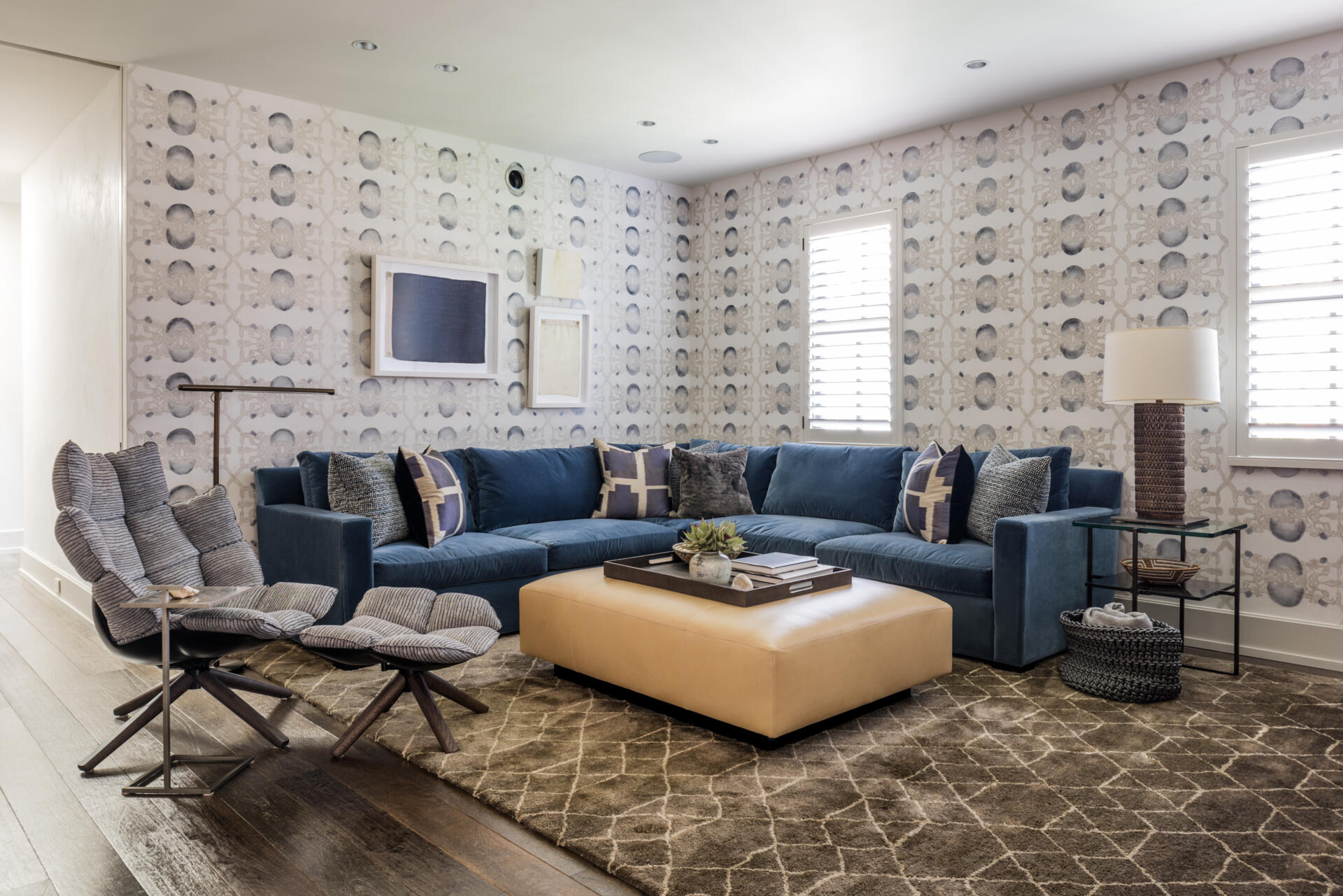 A cozy contemporary media room with a navy-blue sectional sofa, oversized cream ottoman, sculptural lounge chair, blue-gray patterned wallpaper, and textured furnishings.