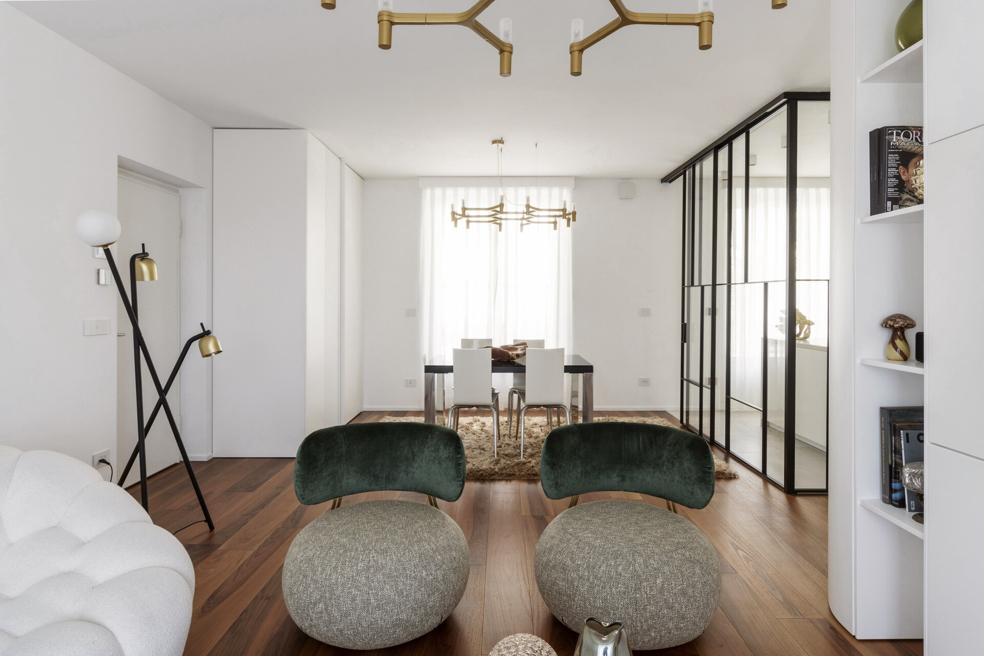 A modern dining area combines a glossy black table, white chairs, a textured rug, and a branching brass chandelier beside a glass-enclosed kitchen.