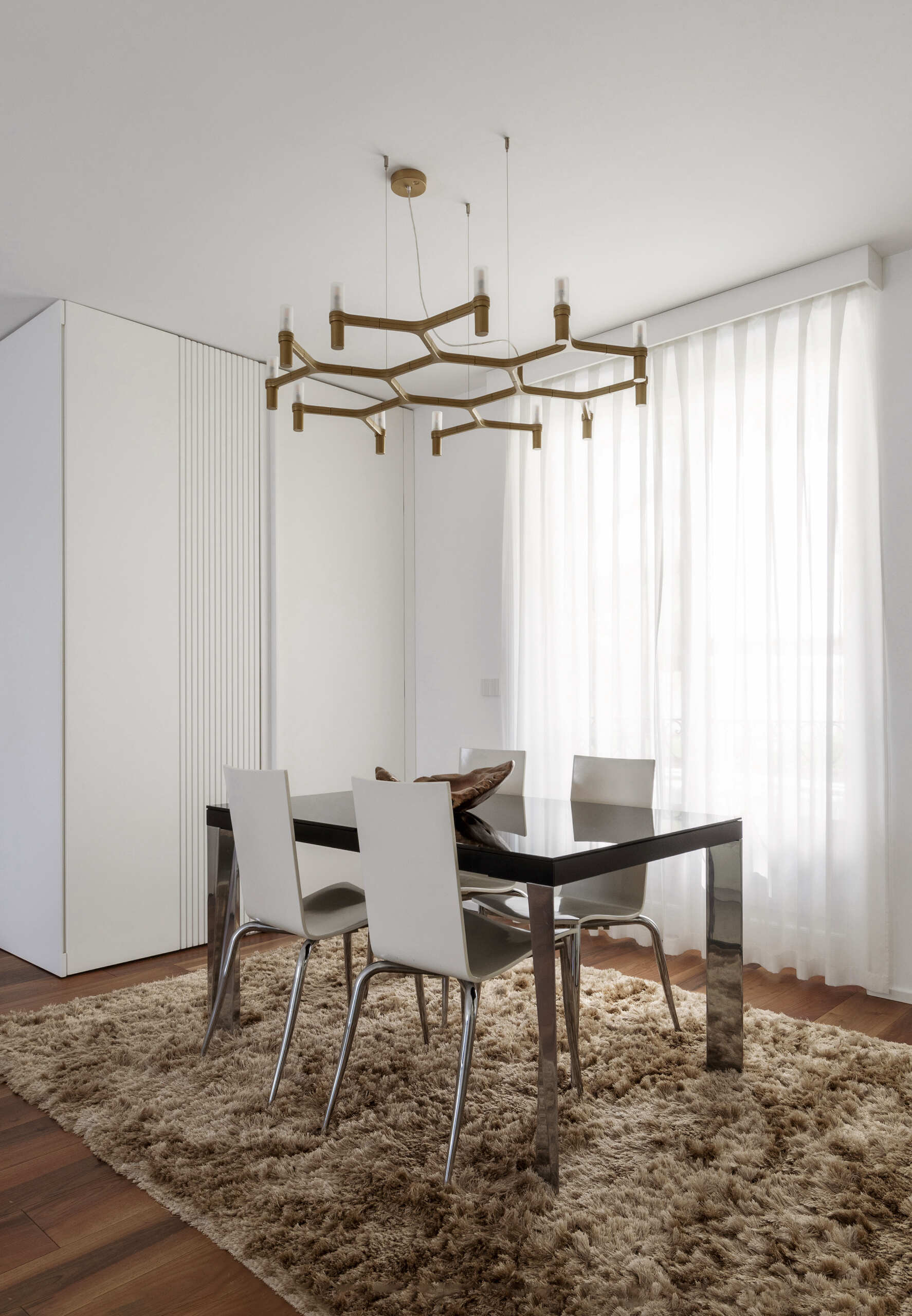A modern dining area combines a glossy black table, white chairs, a textured rug, and a branching brass chandelier beside a glass-enclosed kitchen.
