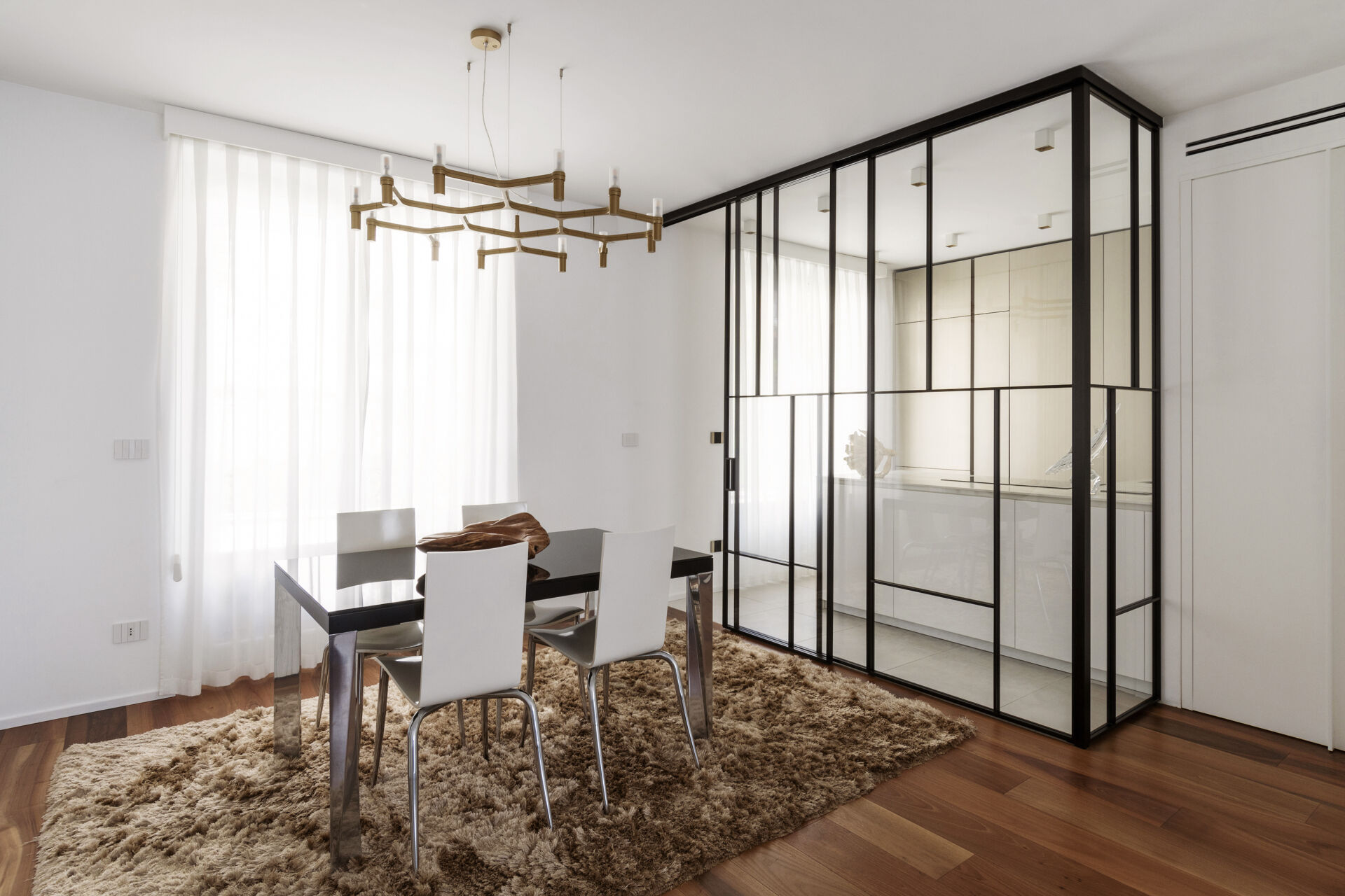 A modern dining area combines a glossy black table, white chairs, a textured rug, and a branching brass chandelier beside a glass-enclosed kitchen.