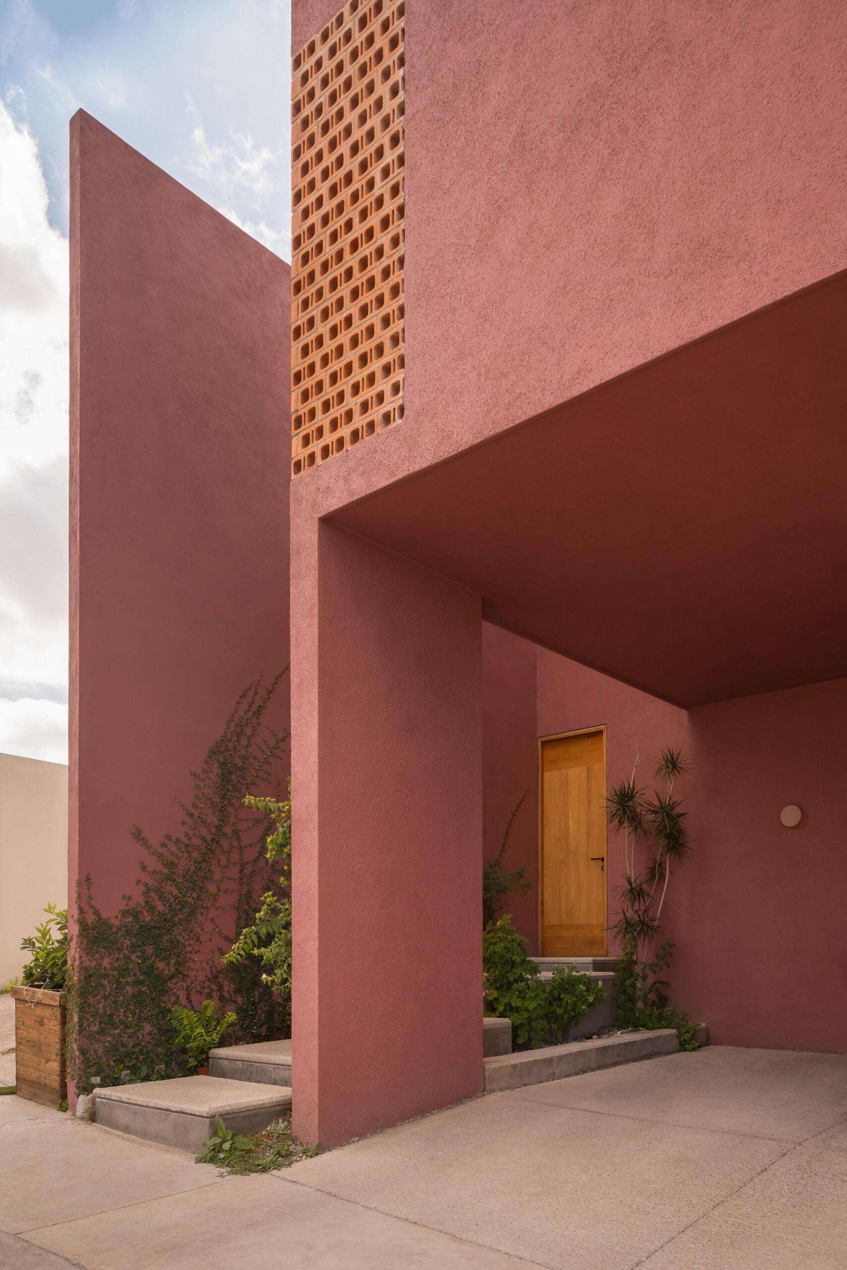 A modern Mexican home features staggered concrete entry steps, pink stucco walls, desert-inspired landscaping, climbing plants, and a natural wood front door.