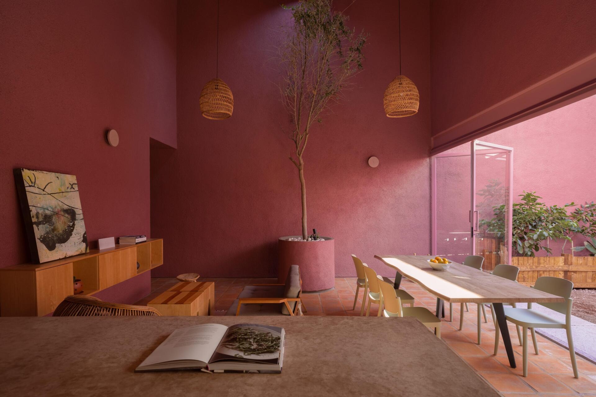 Inside a modern pink house in Mexico, a double-height living room features terracotta floors, a tree beneath a circular skylight, woven pendant lighting, and a wood kitchen with a concrete breakfast bar.