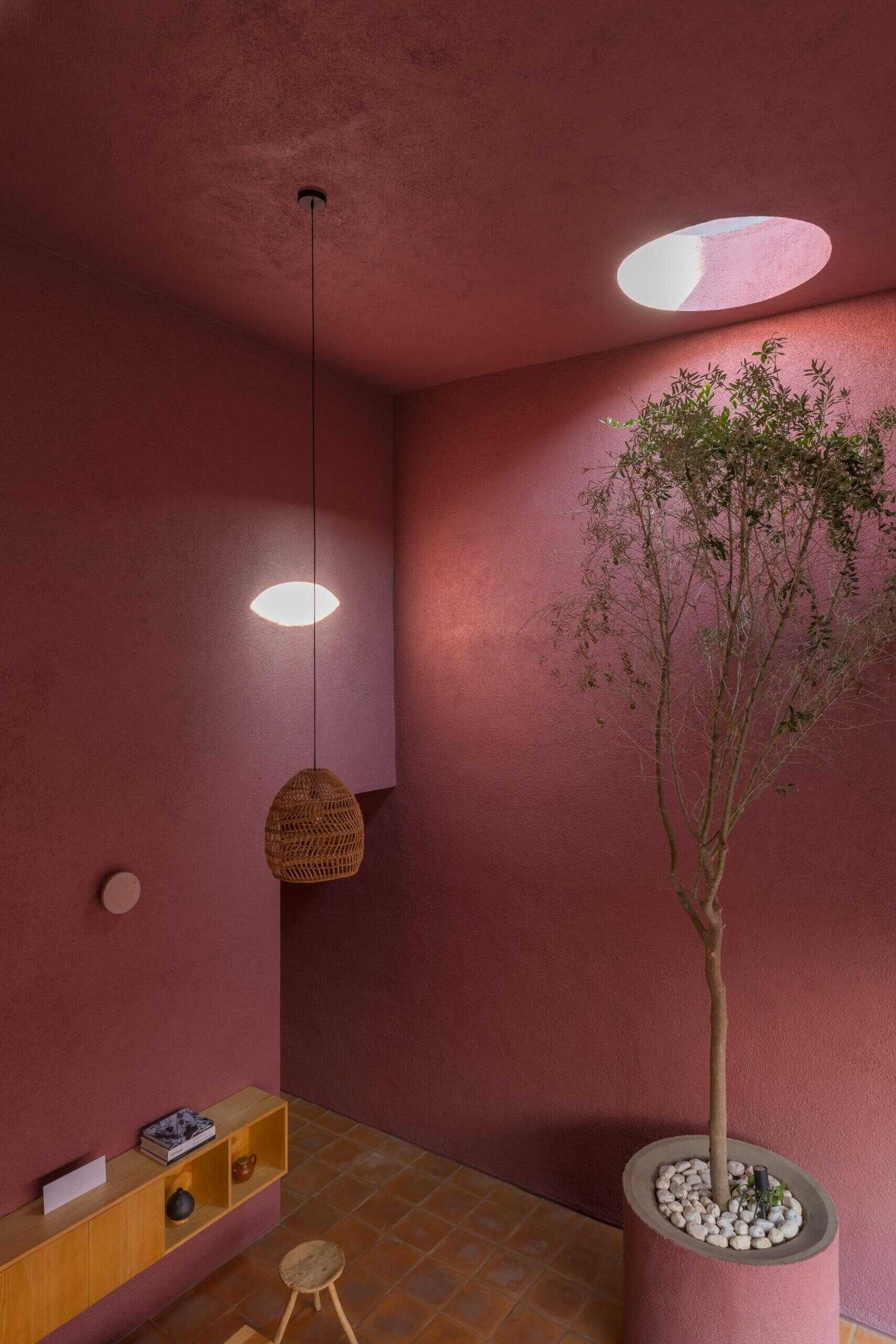 Inside a modern pink house in Mexico, a double-height living room features terracotta floors, a tree beneath a circular skylight, woven pendant lighting, and a wood kitchen with a concrete breakfast bar.