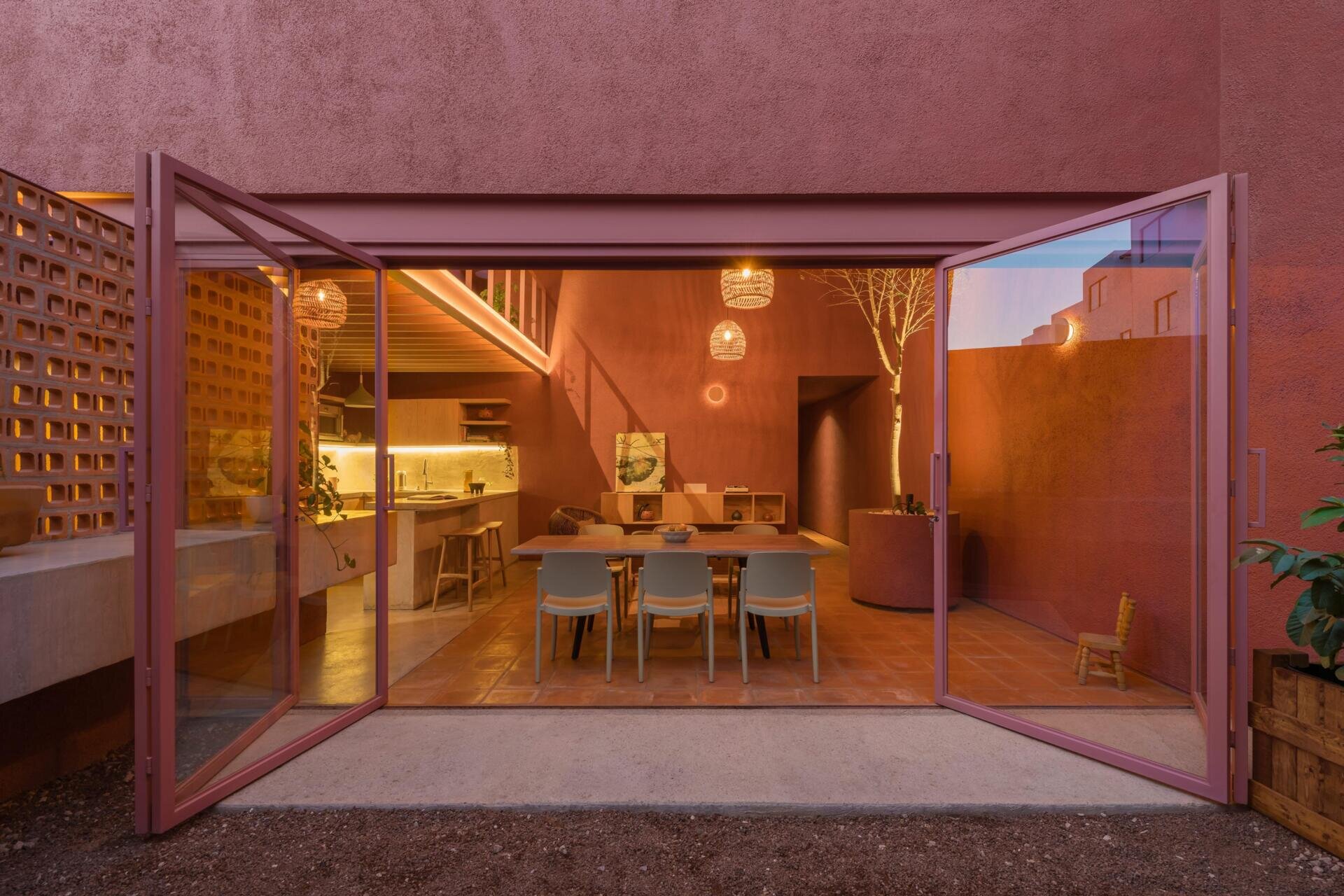 A modern dining room with pink-framed folding glass doors opens to a private courtyard with perforated brick screens, raised garden beds, and pink exterior walls.