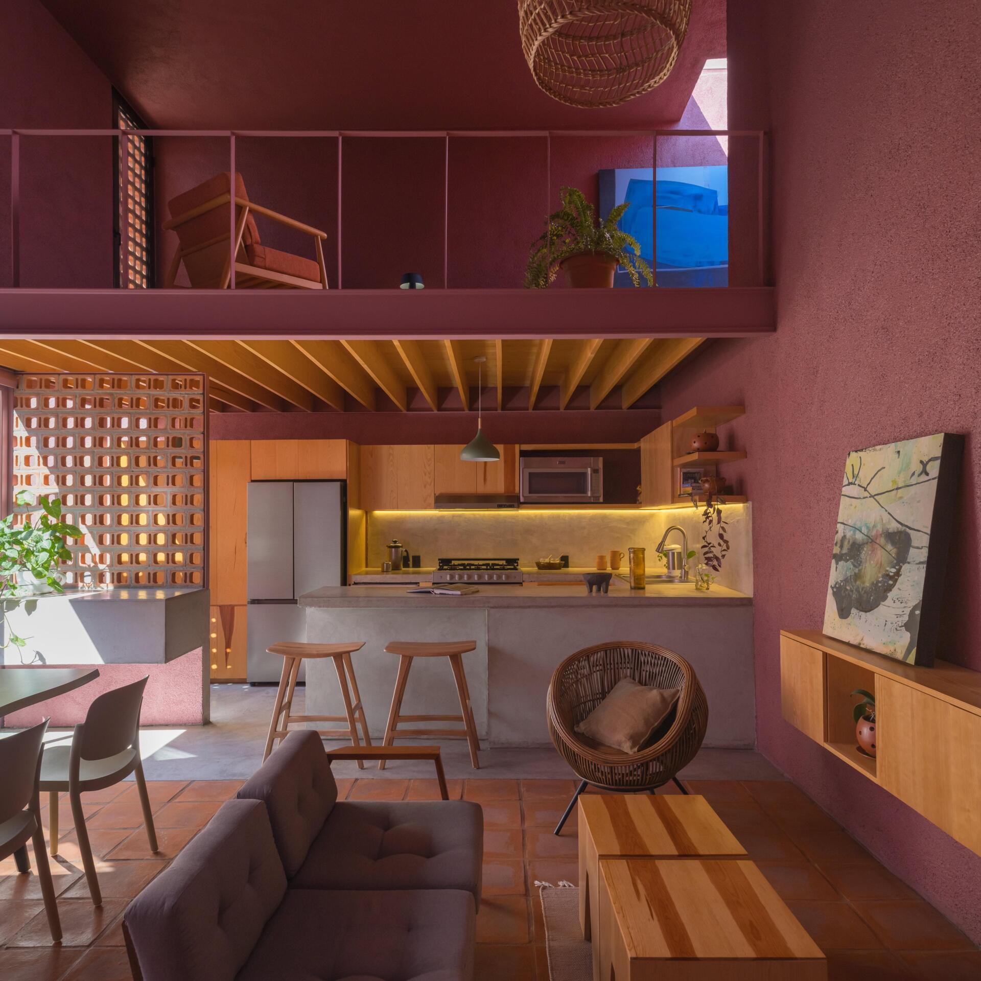 Inside a modern pink house in Mexico, a double-height living room features terracotta floors, a tree beneath a circular skylight, woven pendant lighting, and a wood kitchen with a concrete breakfast bar.