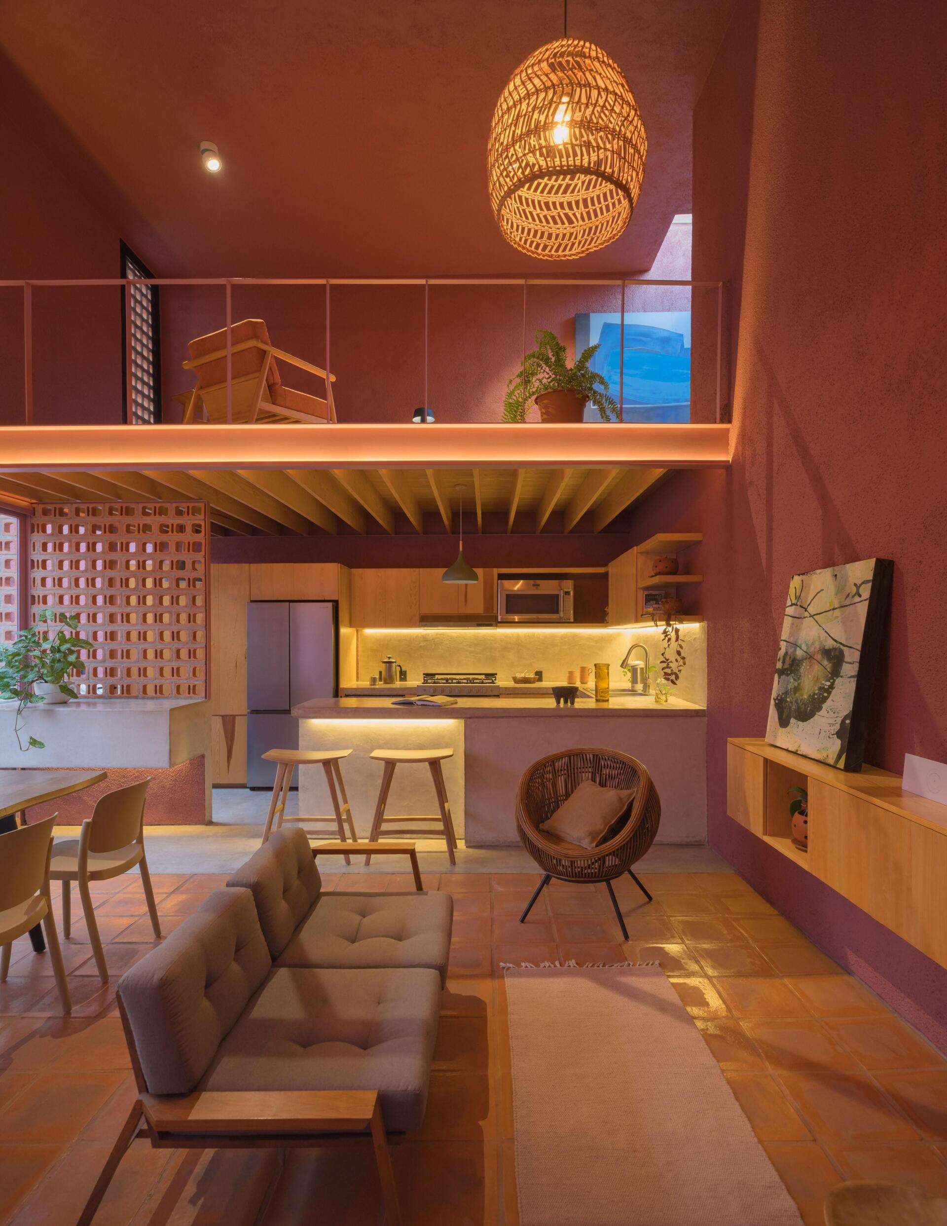 Inside a modern pink house in Mexico, a double-height living room features terracotta floors, a tree beneath a circular skylight, woven pendant lighting, and a wood kitchen with a concrete breakfast bar.