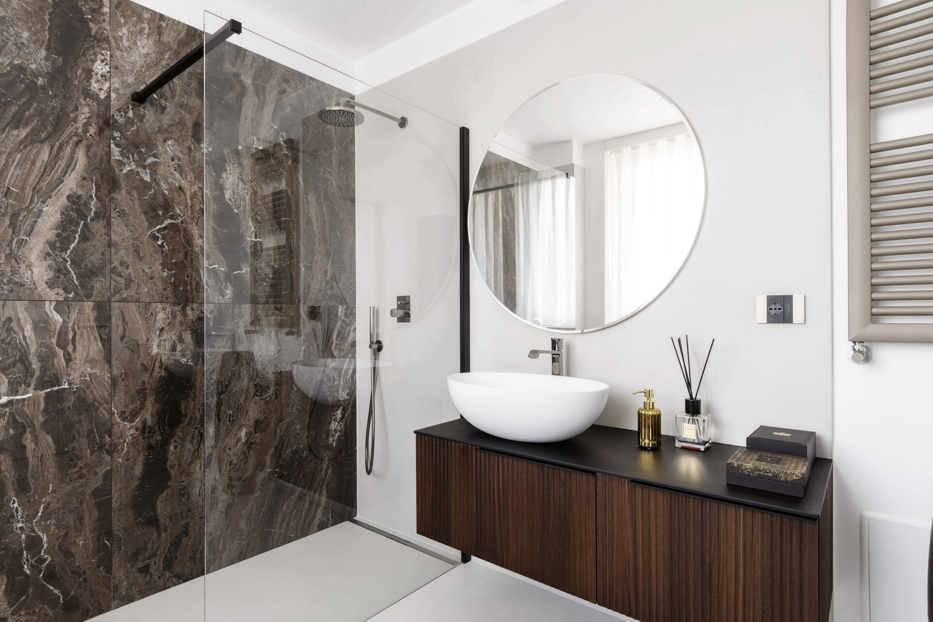 A modern primary bathroom features dramatic marble-look shower tiles, a frameless glass enclosure, a floating dark timber vanity, and a large round mirror.