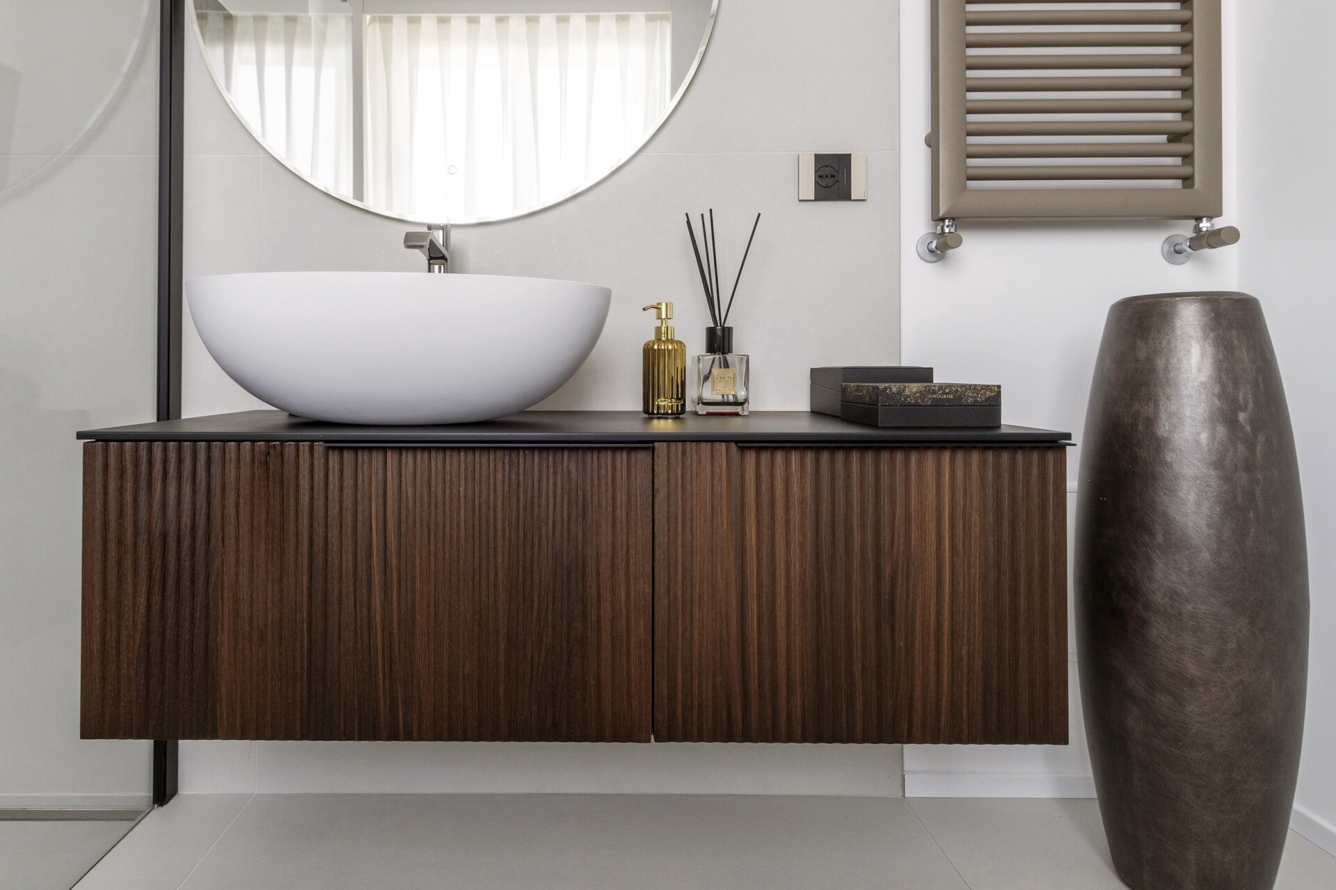 A modern primary bathroom features dramatic marble-look shower tiles, a frameless glass enclosure, a floating dark timber vanity, and a large round mirror.