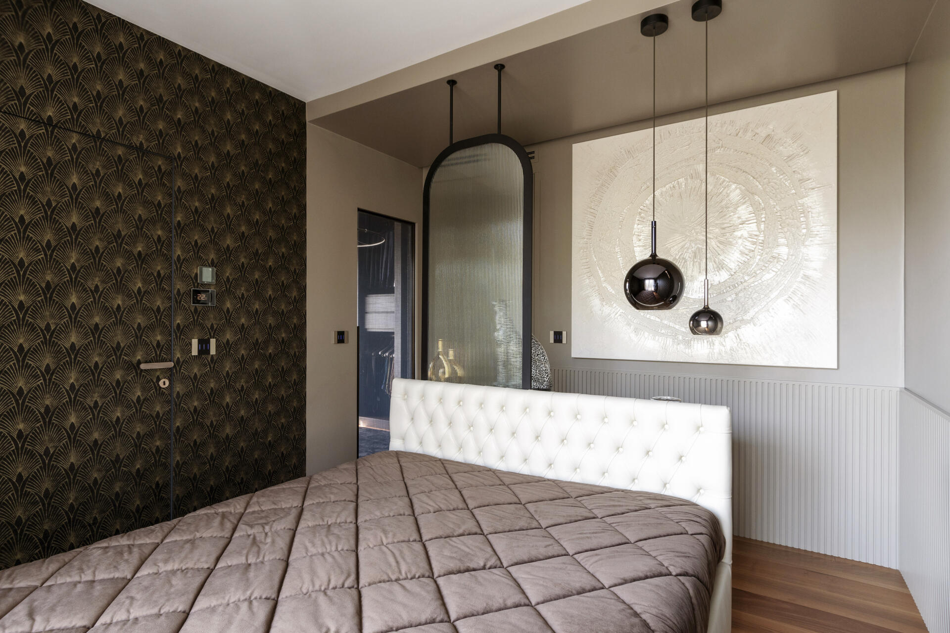 This primary bedroom features Art Deco wallpaper, a white upholstered headboard, black pendant lights, and a custom dressing counter with an arched glass partition.