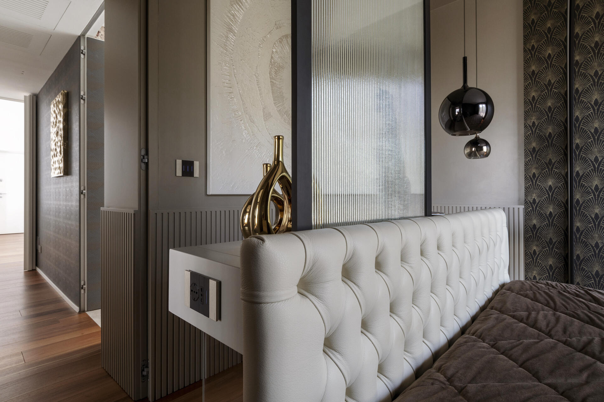 This primary bedroom features Art Deco wallpaper, a white upholstered headboard, black pendant lights, and a custom dressing counter with an arched glass partition.