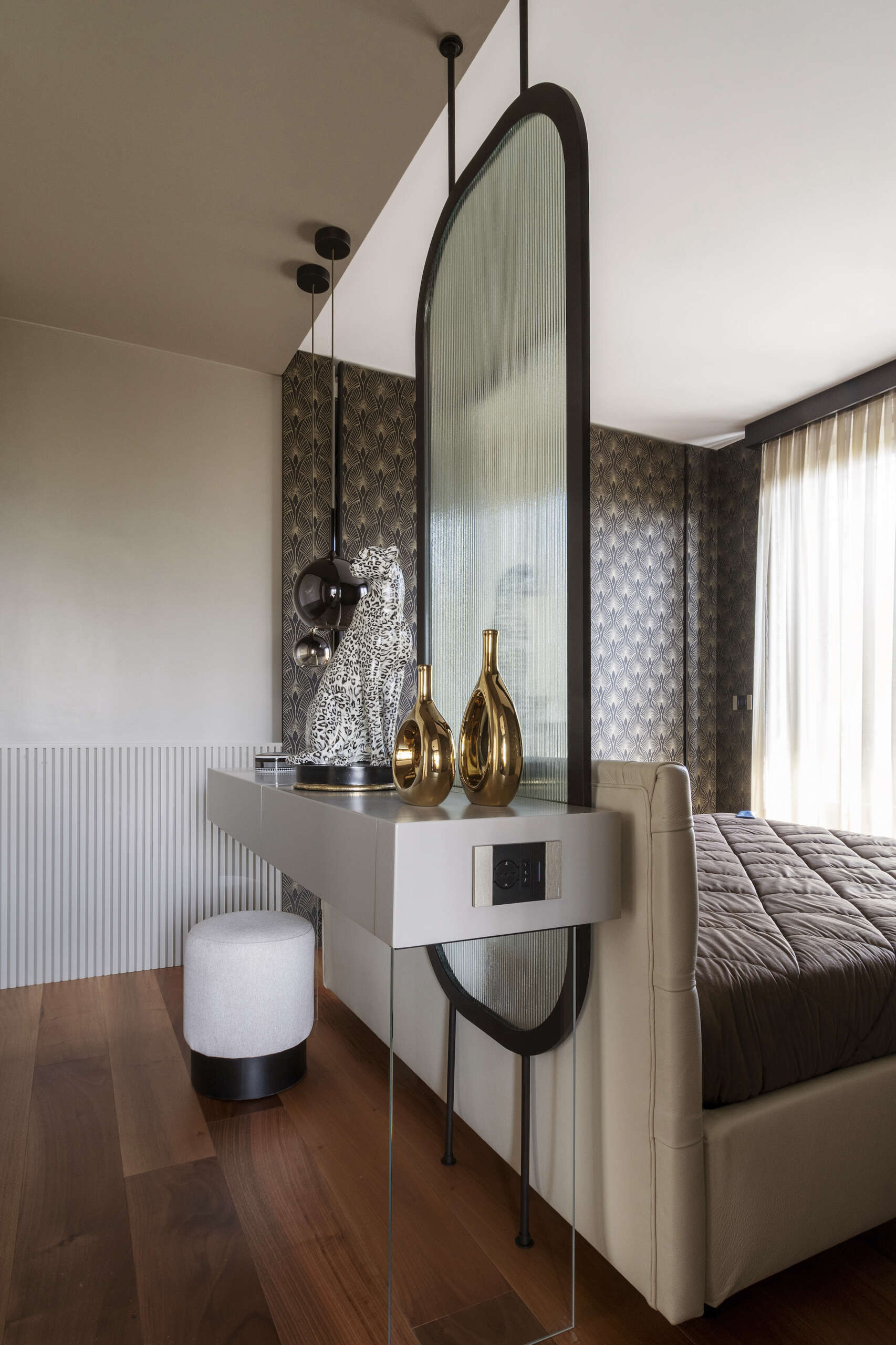 This primary bedroom features Art Deco wallpaper, a white upholstered headboard, black pendant lights, and a custom dressing counter with an arched glass partition.