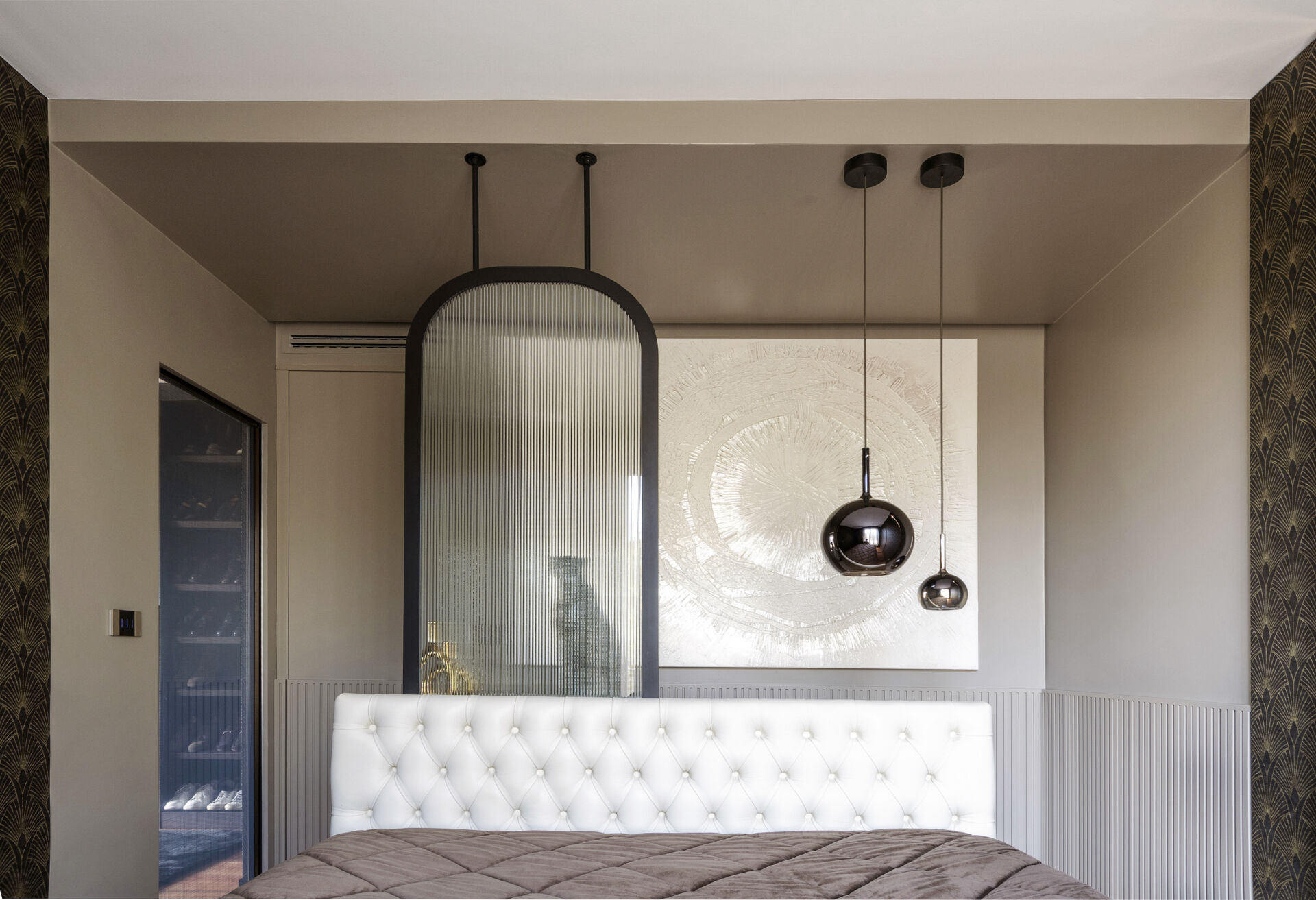 This primary bedroom features Art Deco wallpaper, a white upholstered headboard, black pendant lights, and a custom dressing counter with an arched glass partition.
