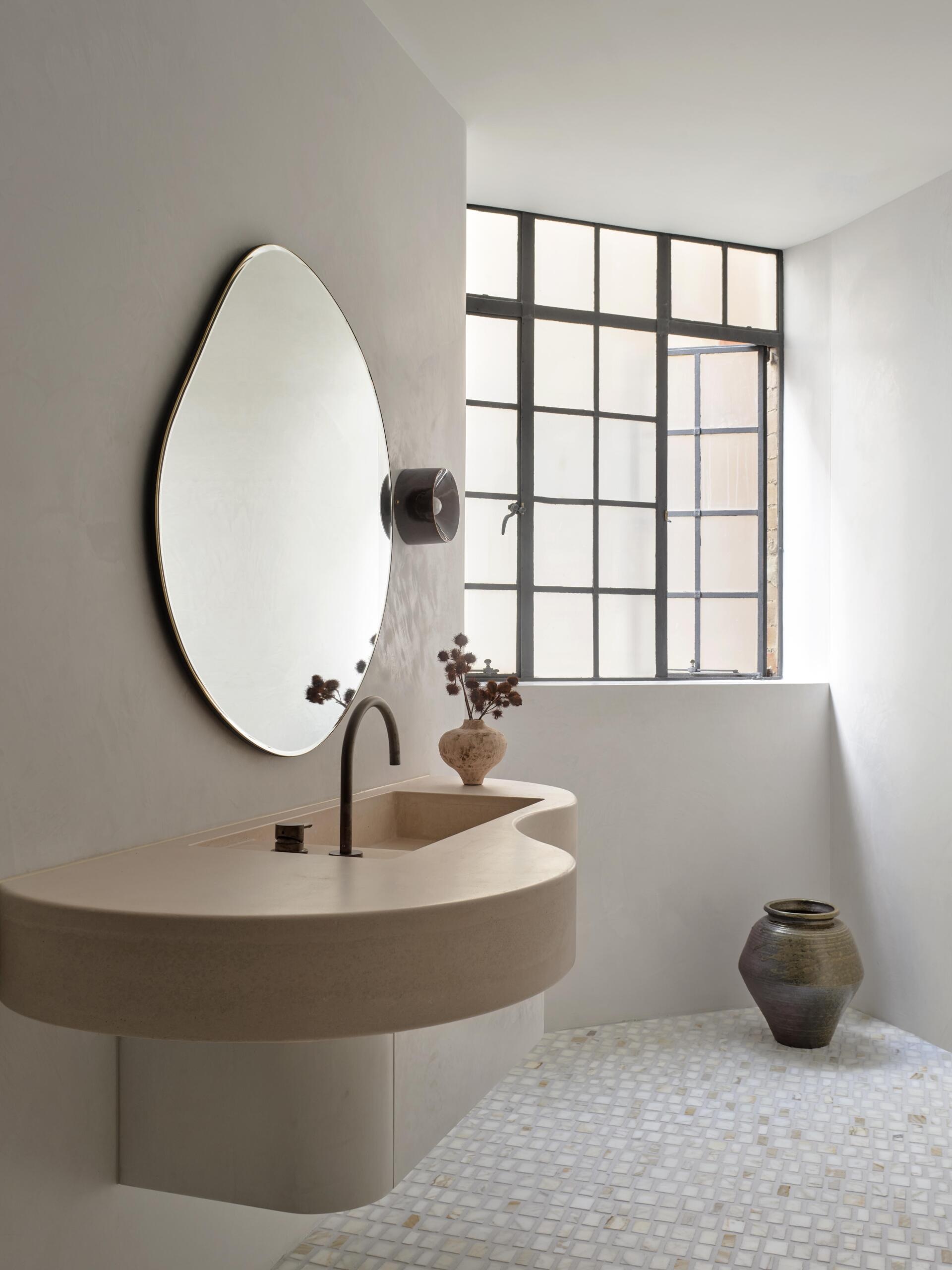A sculptural contemporary bathroom vanity with a curved concrete countertop, integrated rectangular sink, and oversized organic-shaped mirror. Black fixtures, small square floor tiles, and an original steel-framed industrial window complete the minimalist bathroom.