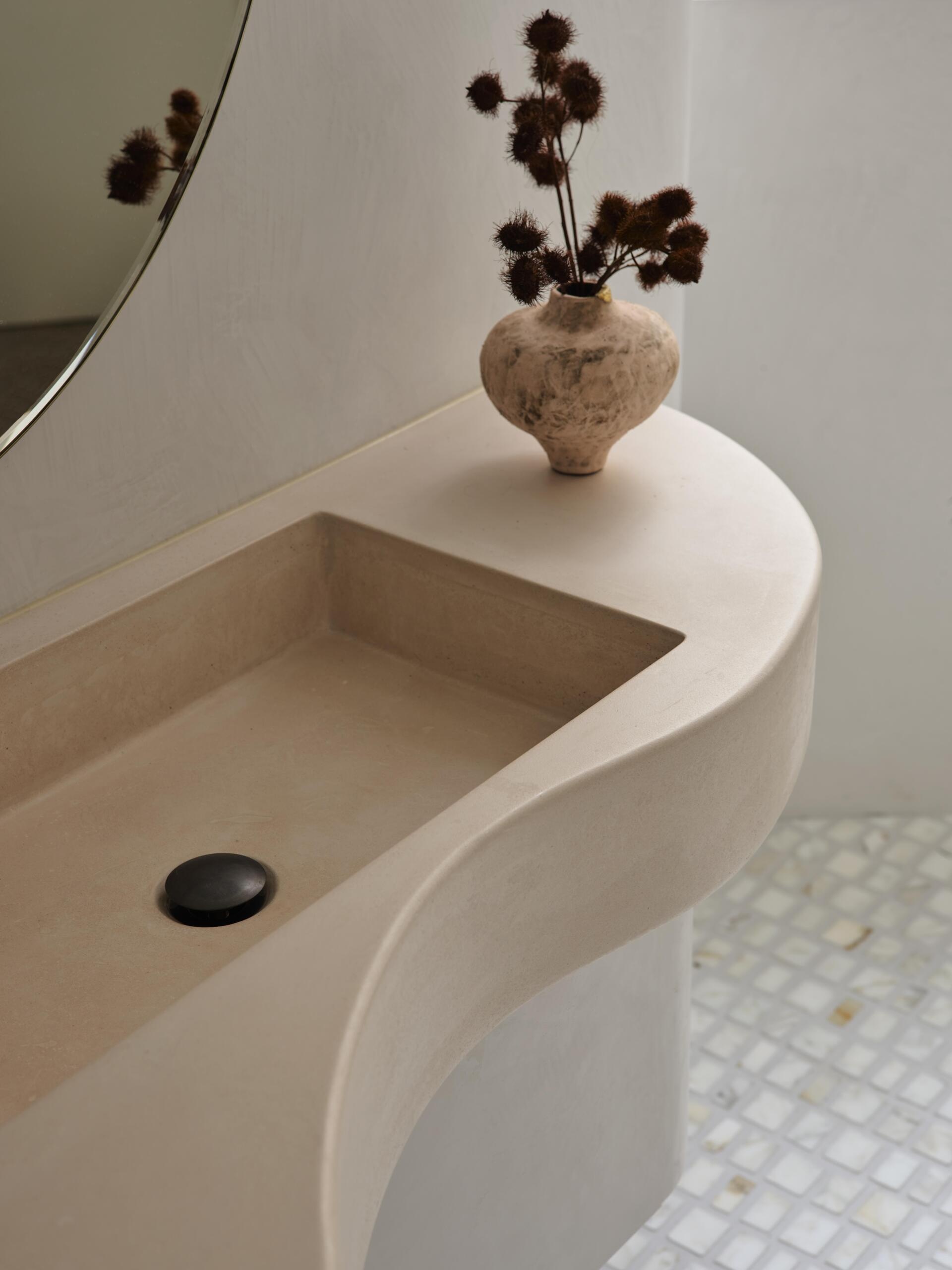 A close-up of a custom concrete bathroom sink with a rectangular basin, rounded edges, and a flowing countertop design. A small ceramic vase and pale mosaic floor tiles complement the soft neutral finish.