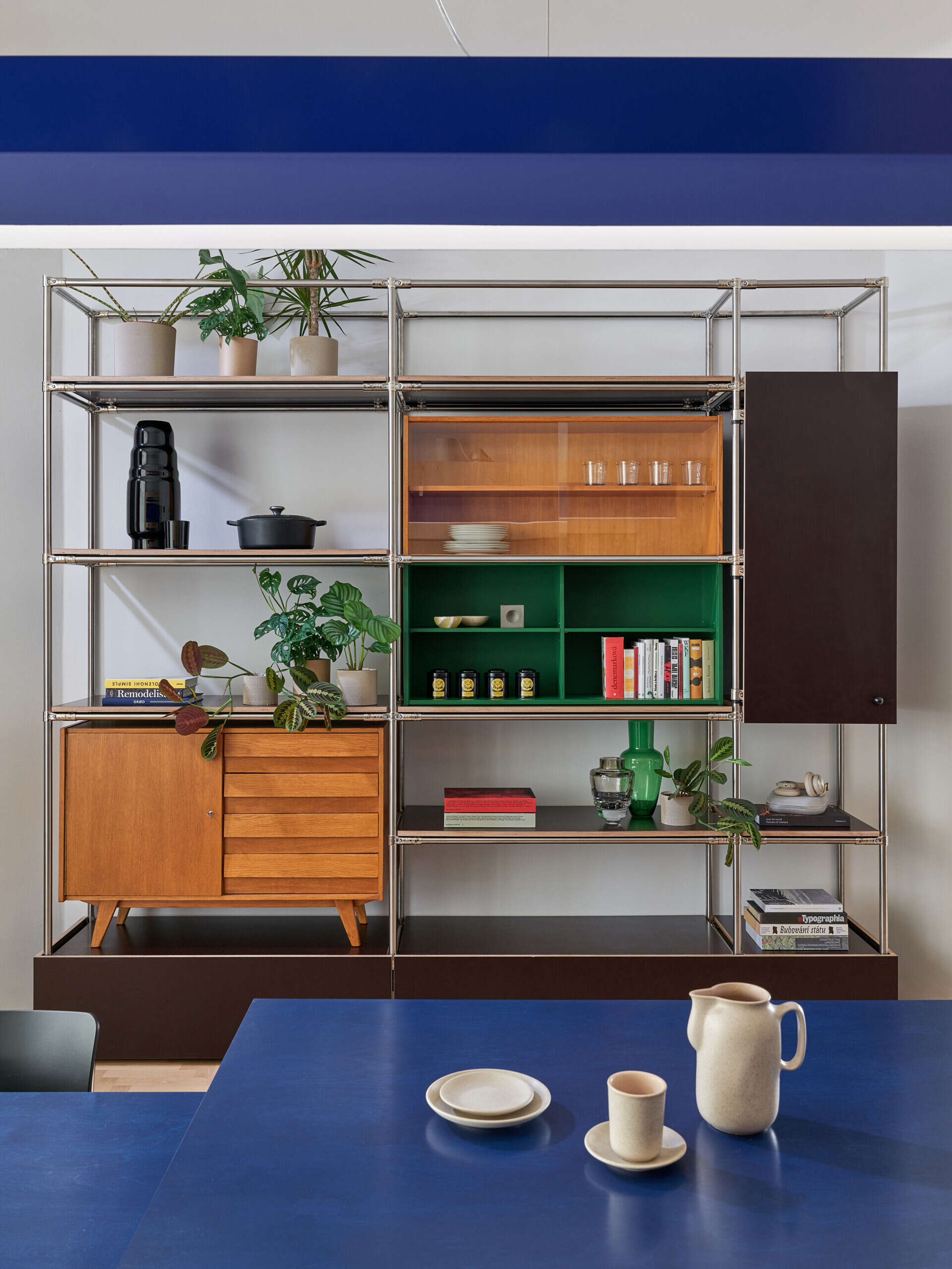 A custom metal shelving system combines open display shelves with warm-toned cabinets, green compartments, plants, and books. The colorful storage wall stands behind a large blue kitchen island.