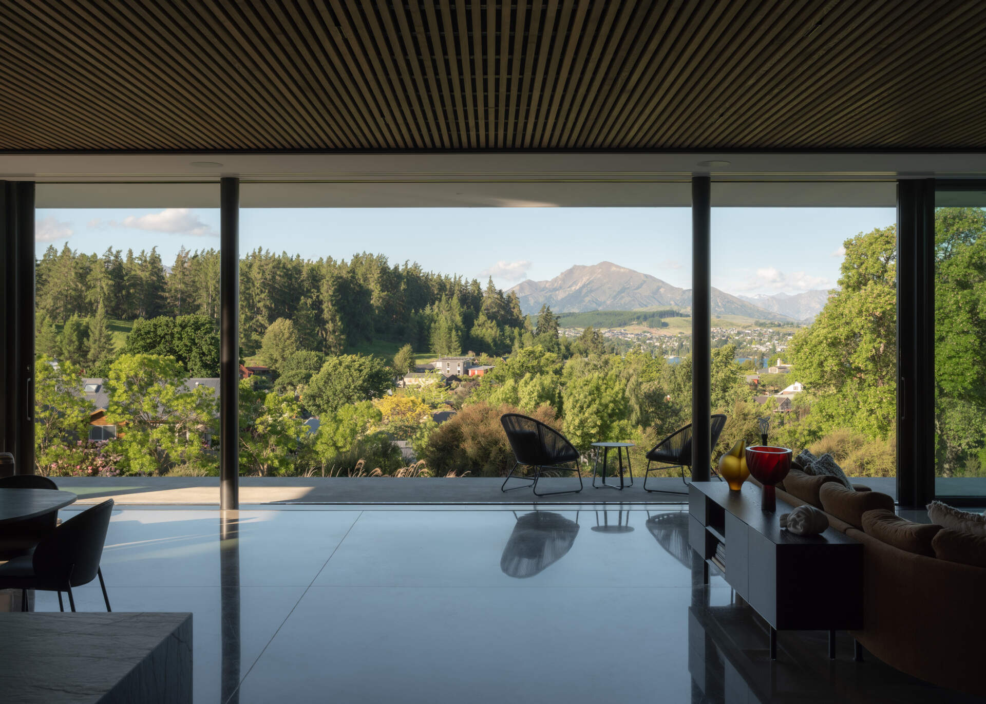 Floor-to-ceiling sliding glass walls open a contemporary living room to a sheltered terrace with mountain views, creating a seamless indoor-outdoor living space in W?naka, New Zealand.