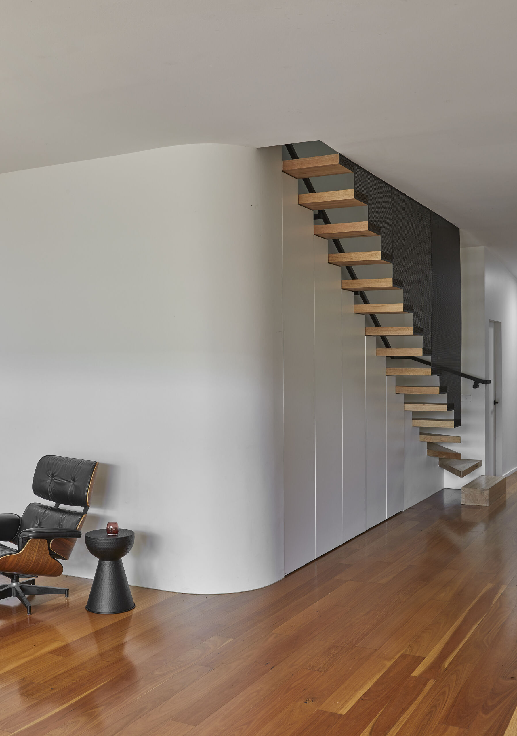 A modern staircase features open timber treads, a black perforated metal screen, and concealed white storage cabinets beneath the steps. A curved wall softens the adjoining living area.