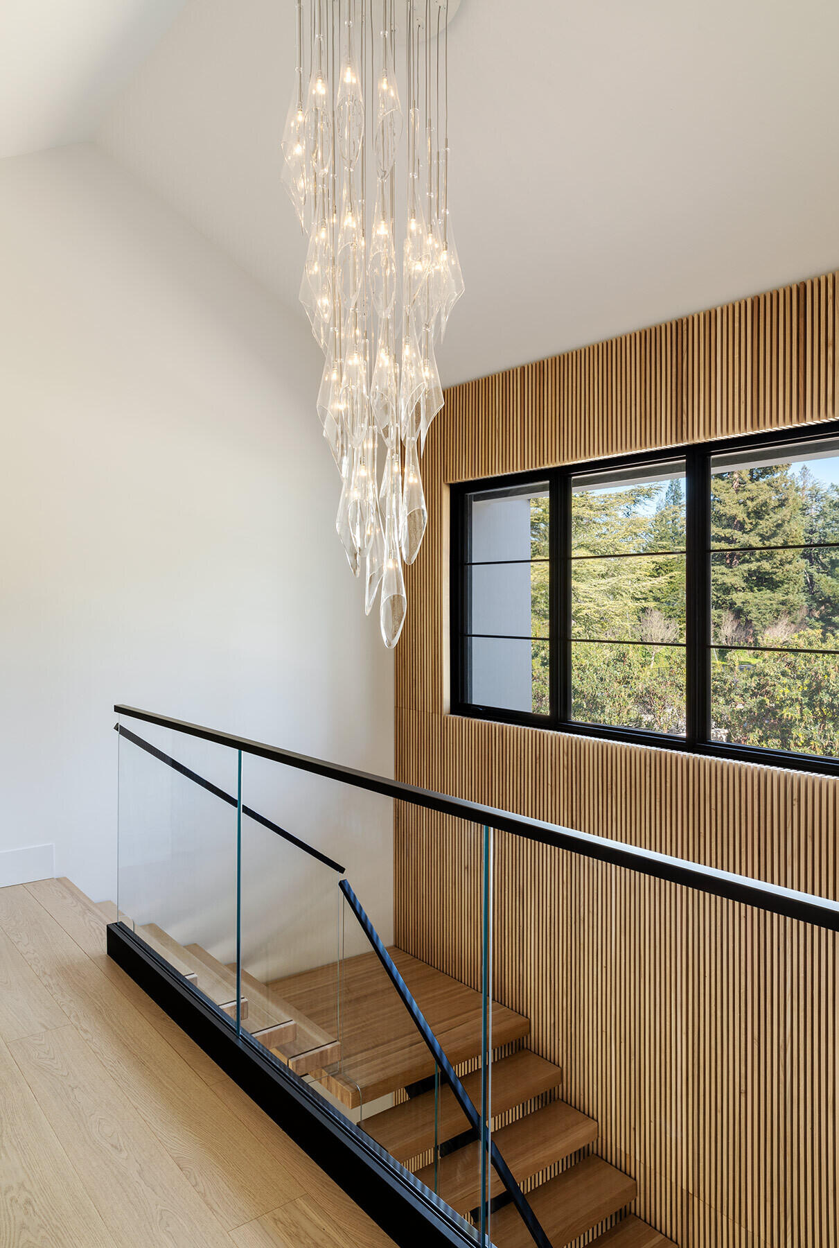 The Atherton Residence staircase pairs pale timber treads, glass balustrades, black handrails and a tall slatted timber wall with a cascading glass light fixture in the double-height stairwell.